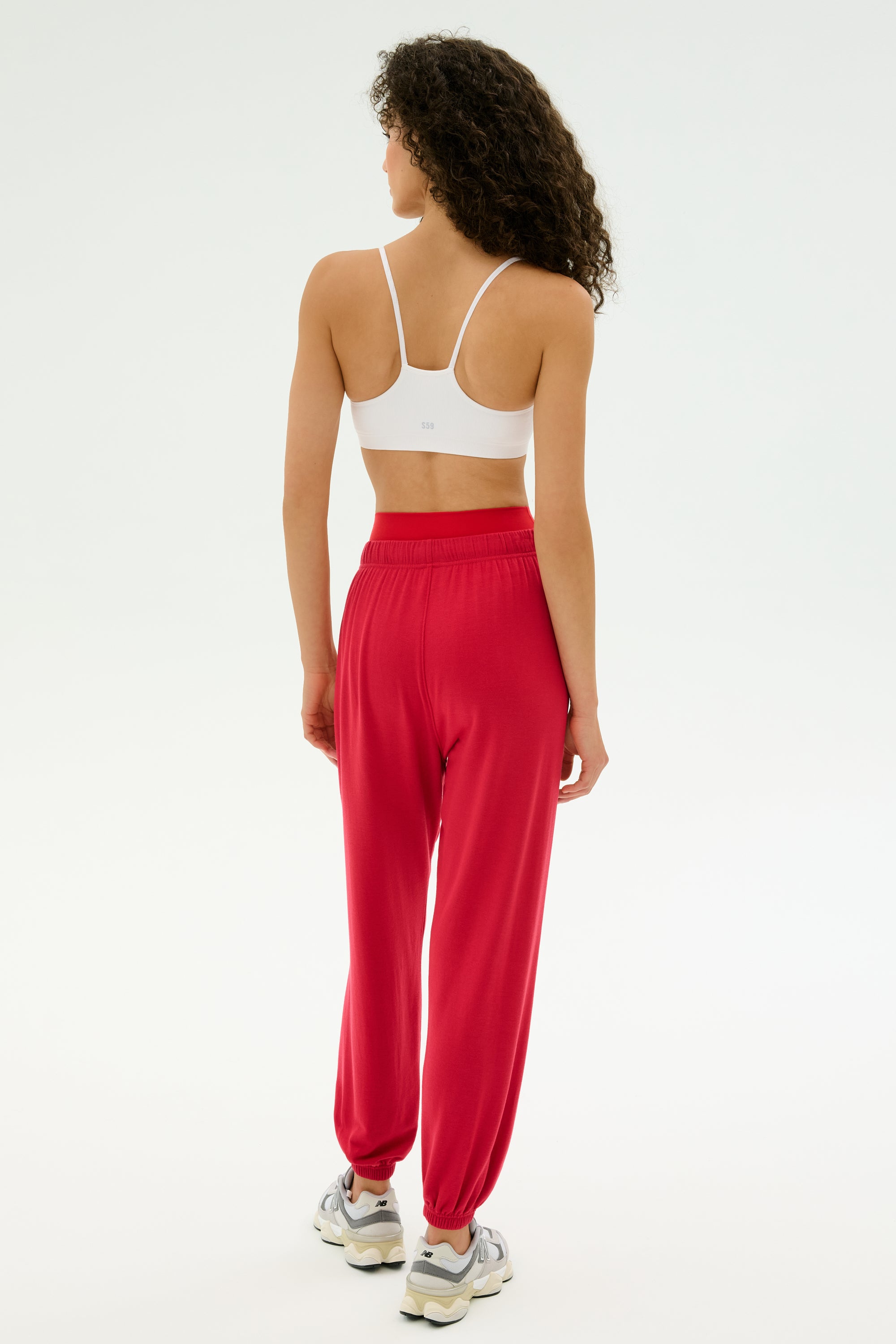 A person faces away, wearing a white sports bra and SPLITS59 Andie Oversized Fleece Sweatpant in Pirate Red, perfect for cool weather workouts or warm-ups, with white sneakers against a plain light background.