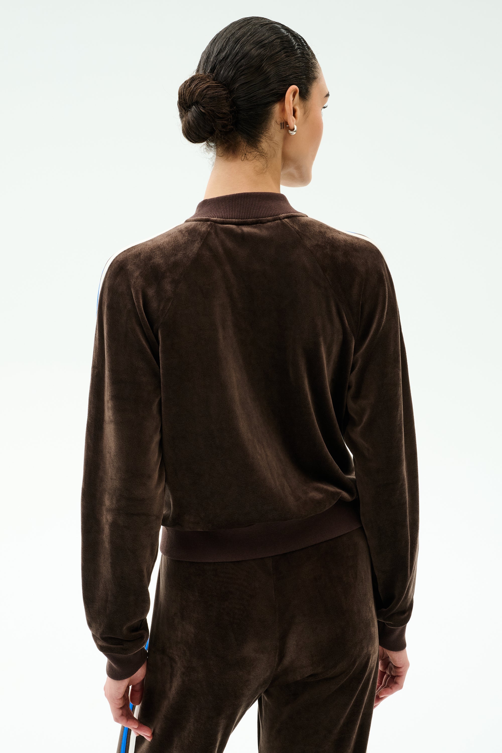 A person with dark hair in a bun is shown from the back, warming up in the SPLITS59 Heritage Velour Bomber - Dark Chocolate/Classic Blue against a plain white background.