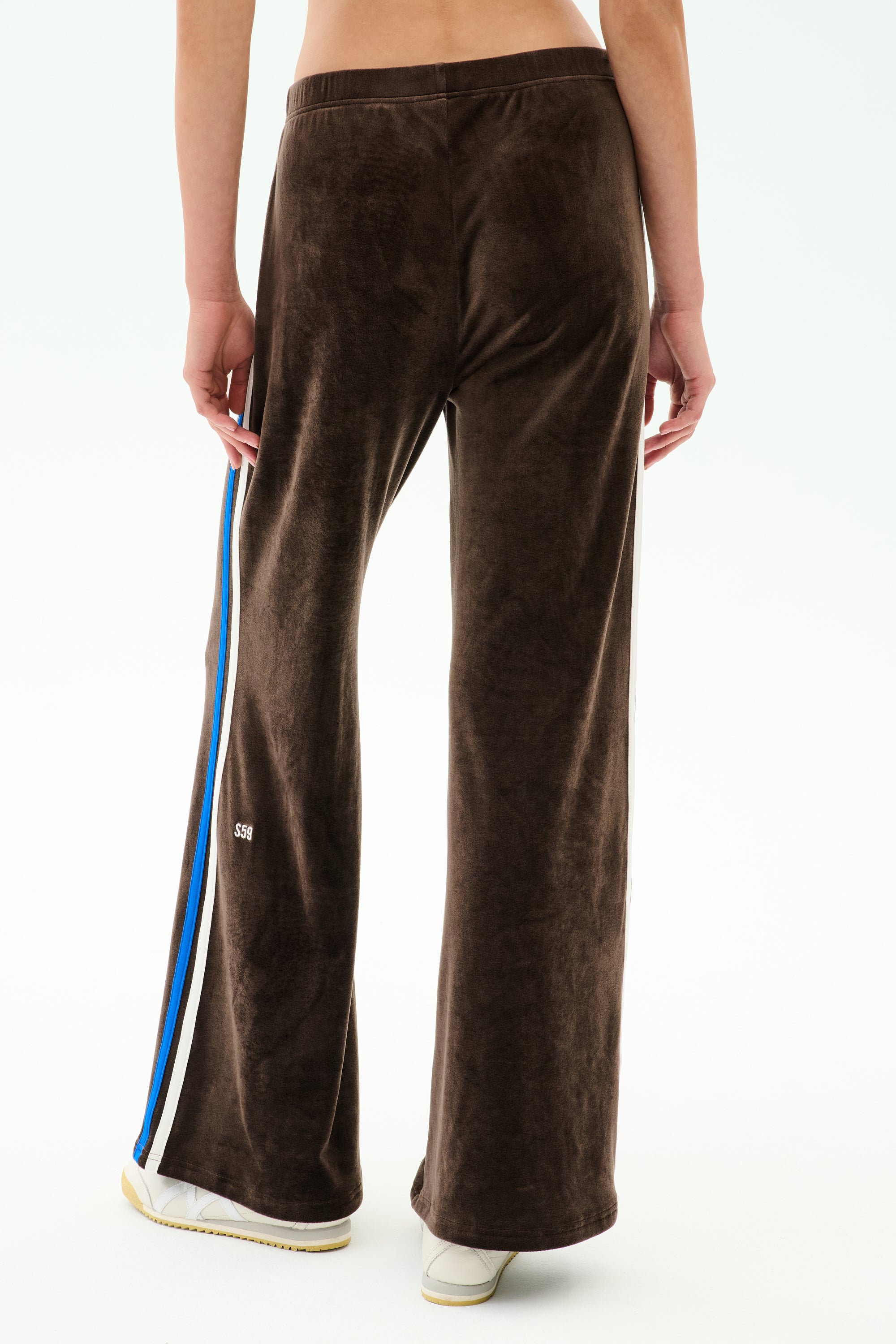 A person models the SPLITS59 Heritage Velour Pant in Dark Chocolate/Classic Blue—ankle-length, wide-leg silhouette with blue and white side stripes—paired with white sneakers, shown from the back against a plain background.