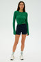 A person wearing a green long-sleeve shirt tied at the waist, SPLITS59 Ella Airweight High Waist Short 3.5" - Indigo/White, white sneakers, and striped socks stands against a plain background.