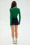 A woman with long brown hair stands facing away, wearing the Arugula-colored Louise Rib Long Sleeve by SPLITS59, black shorts, white socks, and white sneakers against a plain background.