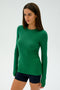 A woman wearing the SPLITS59 Louise Rib Long Sleeve in Arugula and dark spandex shorts stands against a plain background, looking to the side.