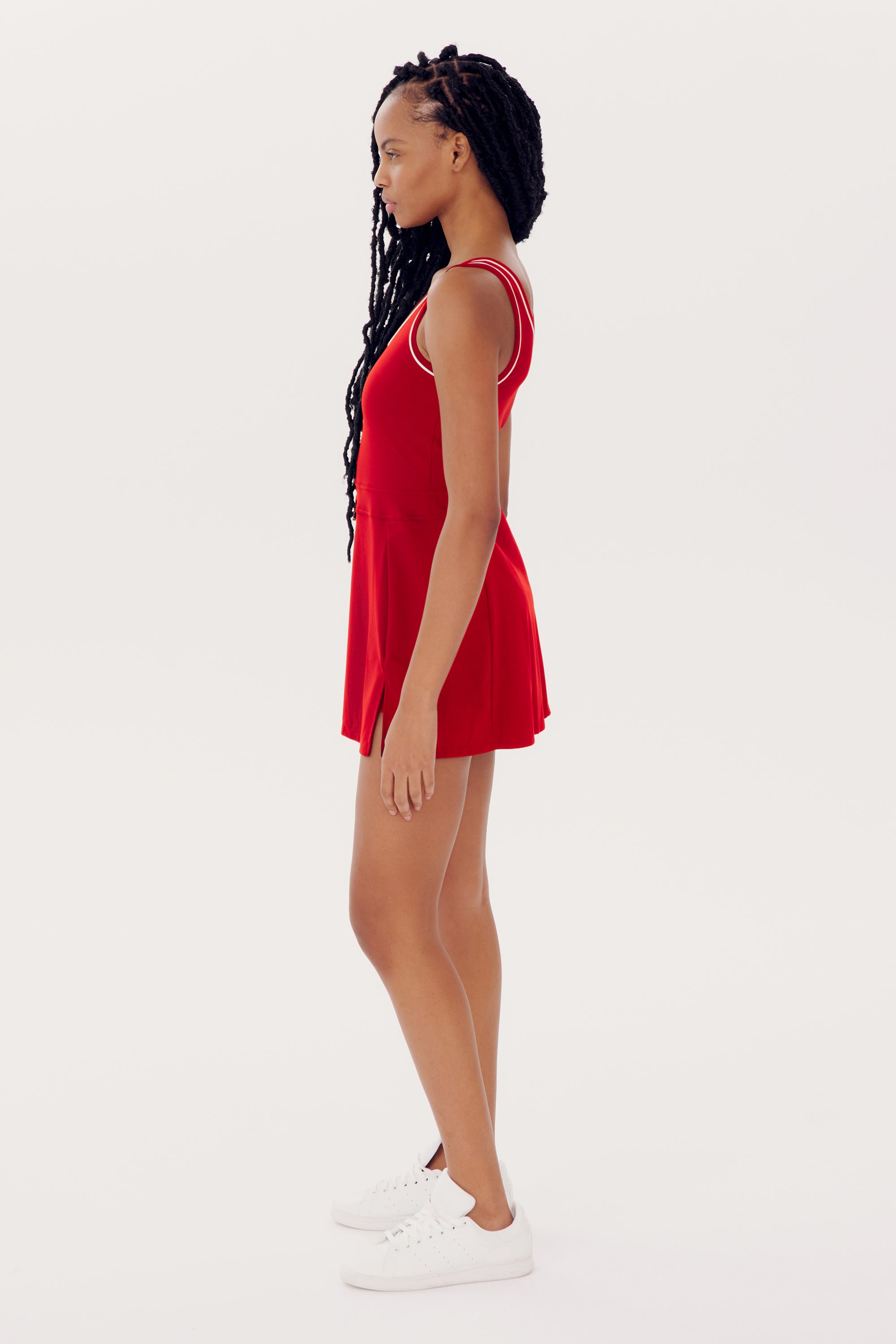 A person with long braided hair is standing in profile view, wearing a sleeveless red SPLITS59 Martina Rigor Dress w/Piping - Pirate Red and white sneakers against a plain white background.