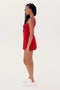 A person with long braided hair is standing in profile view, wearing a sleeveless red SPLITS59 Martina Rigor Dress w/Piping - Pirate Red and white sneakers against a plain white background.