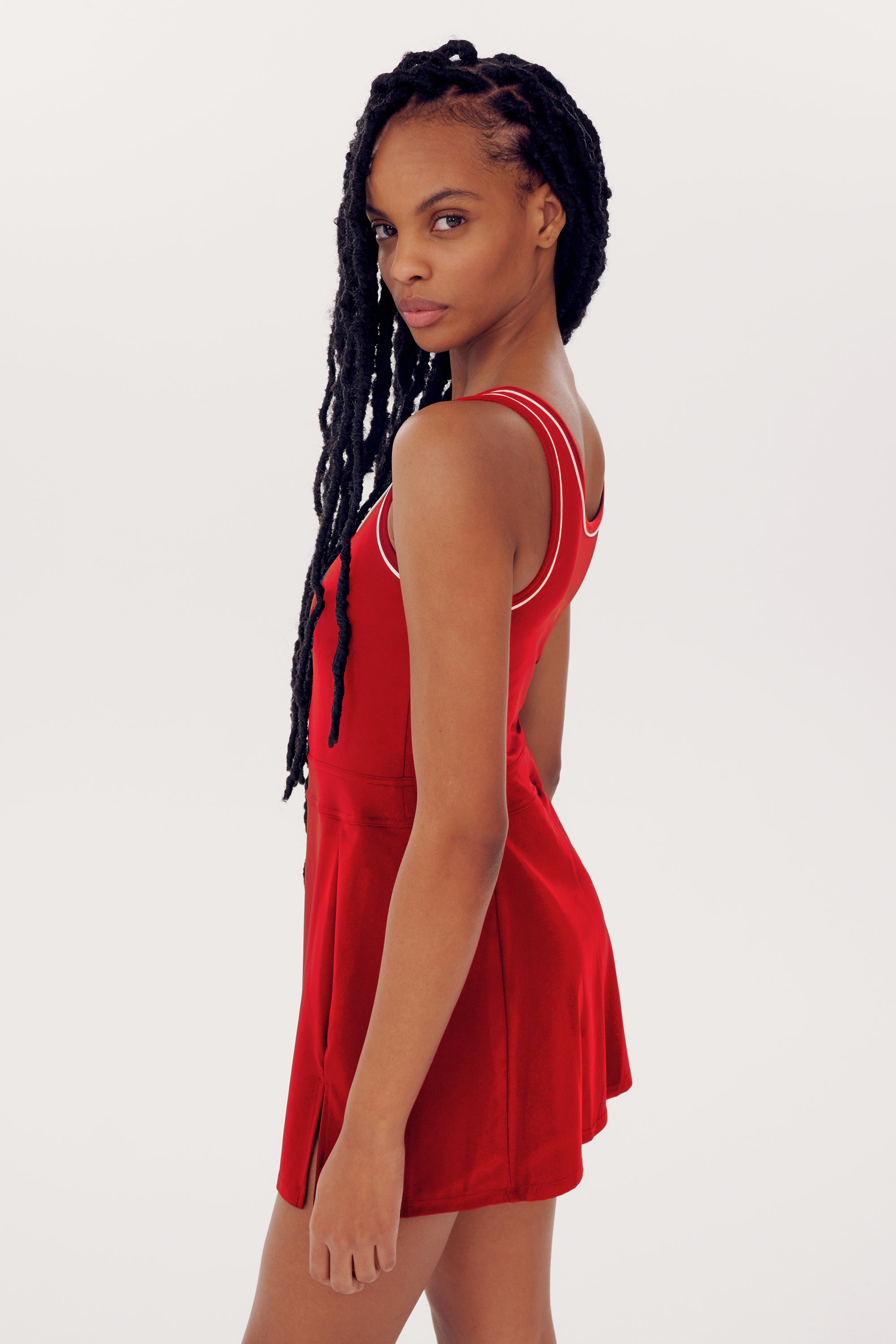 Woman with long braids in a red sleeveless Martina Rigor Dress w/Piping - Pirate Red, made by SPLITS59, standing against a white background, looking over her shoulder.