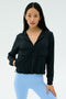A woman faces forward against a plain light background, wearing the SPLITS59 Megan Woven Nylon Jacket in black and light blue spandex leggings.