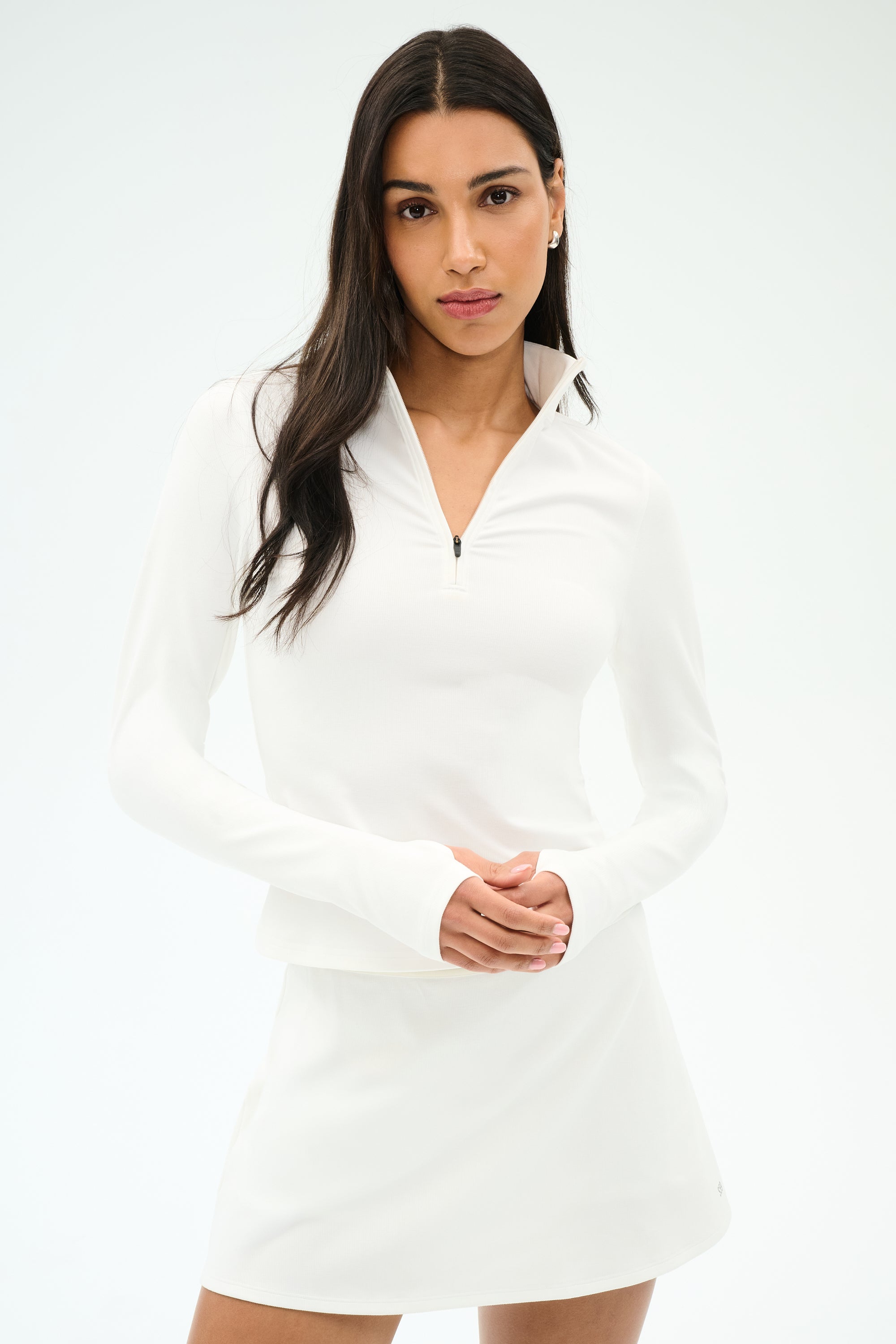 A woman stands against a plain background in the SPLITS59 Stevie Active Rib Half Zip Waist Length top in white, paired with a matching white skirt, her hands loosely clasped in front of her.