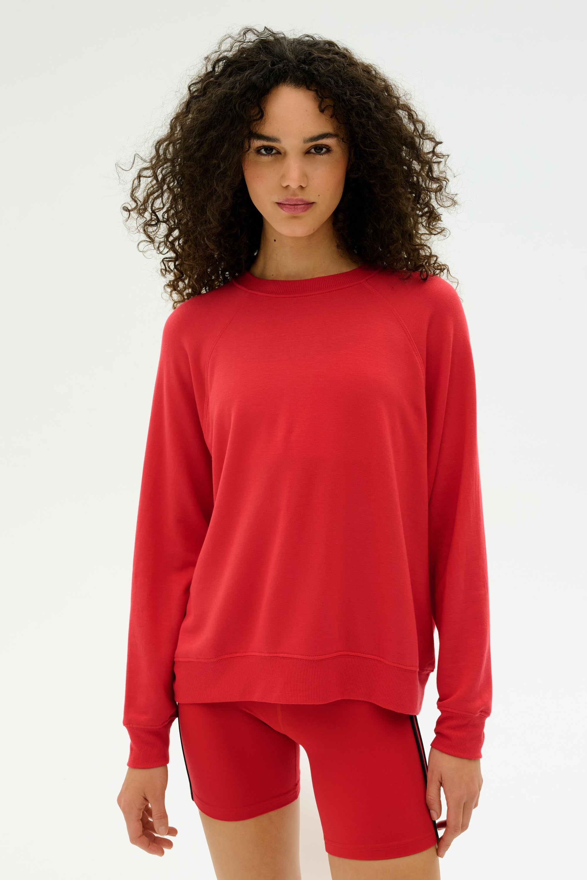 A woman with curly hair wears the SPLITS59 Andie Fleece Sweatshirt in Pirate Red, along with matching shorts, made from stretch modal fabric. She stands against a plain white background. Made in Los Angeles.