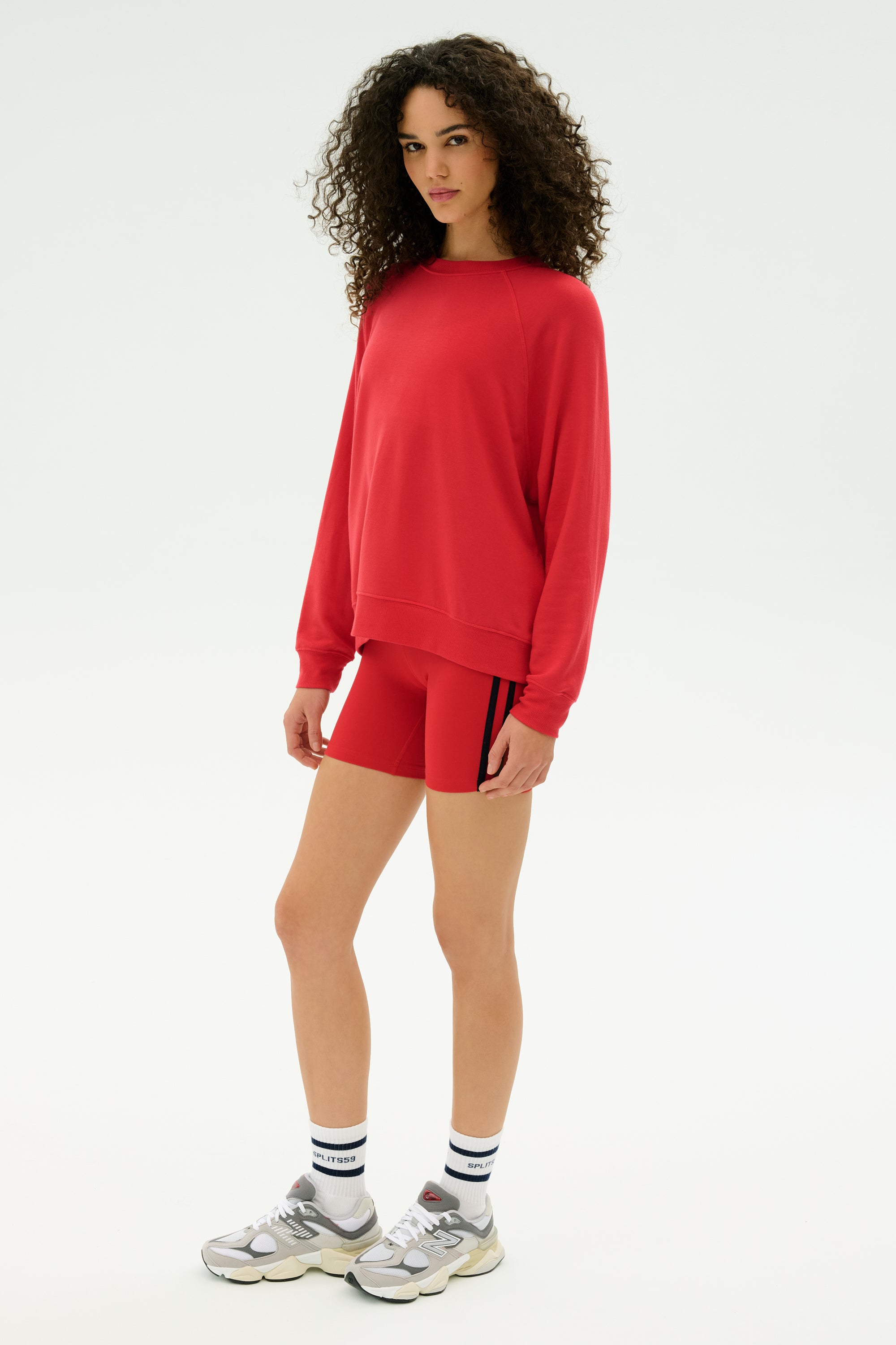 A person stands facing slightly left, wearing the SPLITS59 Andie Fleece Sweatshirt in Pirate Red, matching red shorts, white socks with black stripes, and silver sneakers against a plain white background.