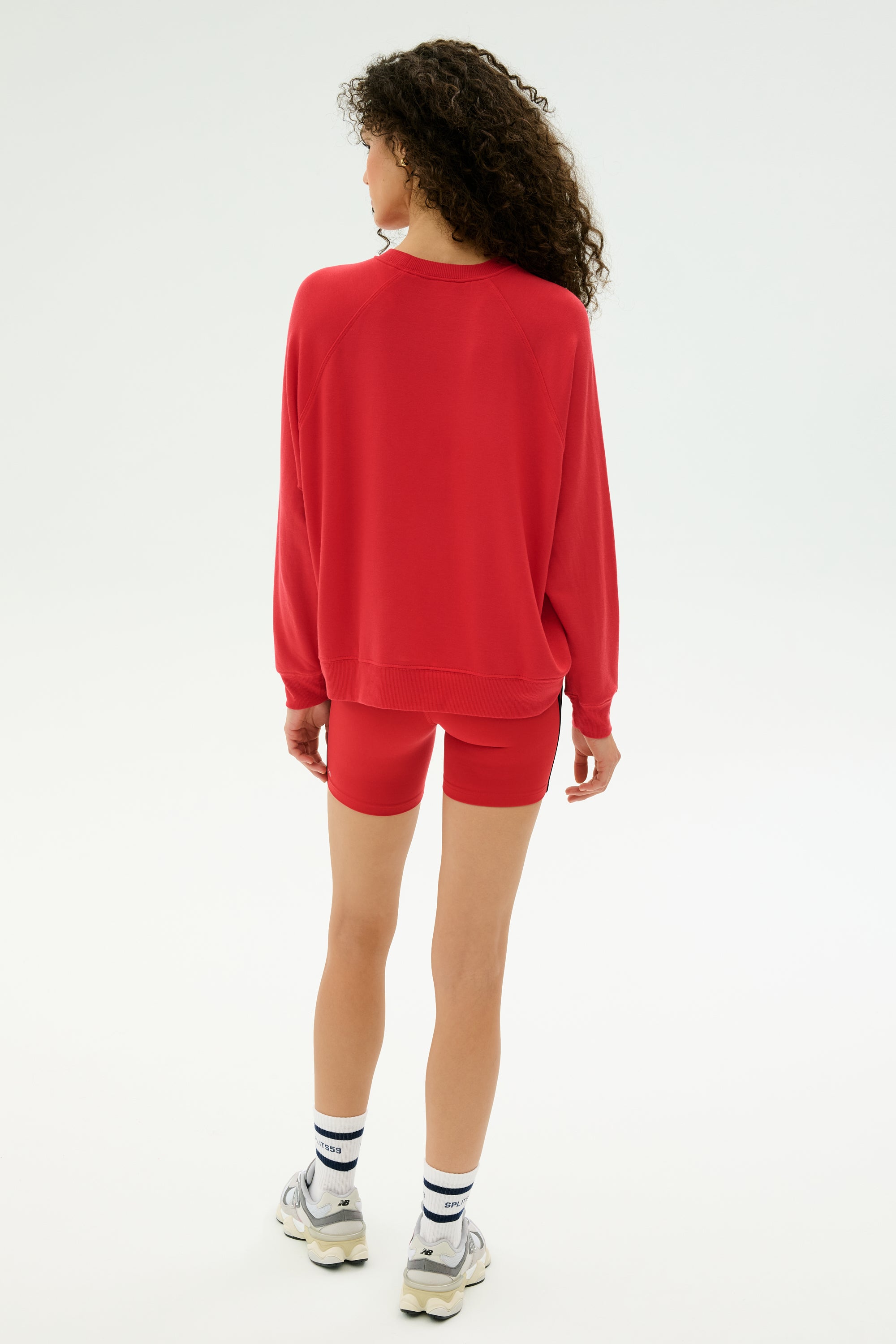 A person with curly hair faces away, wearing the SPLITS59 Andie Fleece Sweatshirt in Pirate Red, matching red shorts, white socks, and white sneakers against a plain light background.