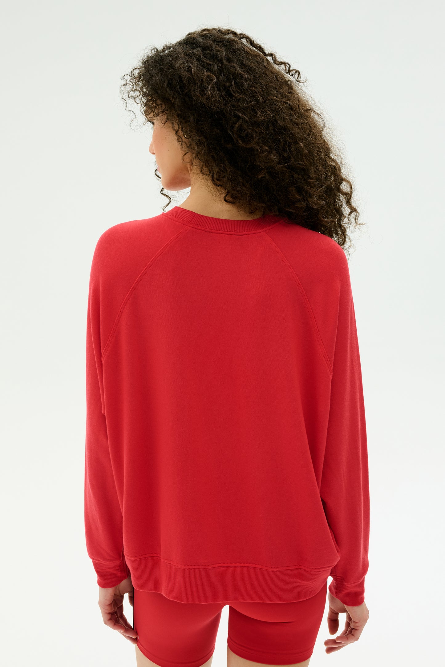 A person with curly hair is shown from the back wearing the SPLITS59 Andie Fleece Sweatshirt in Pirate Red, paired with red shorts, against a plain light background.
