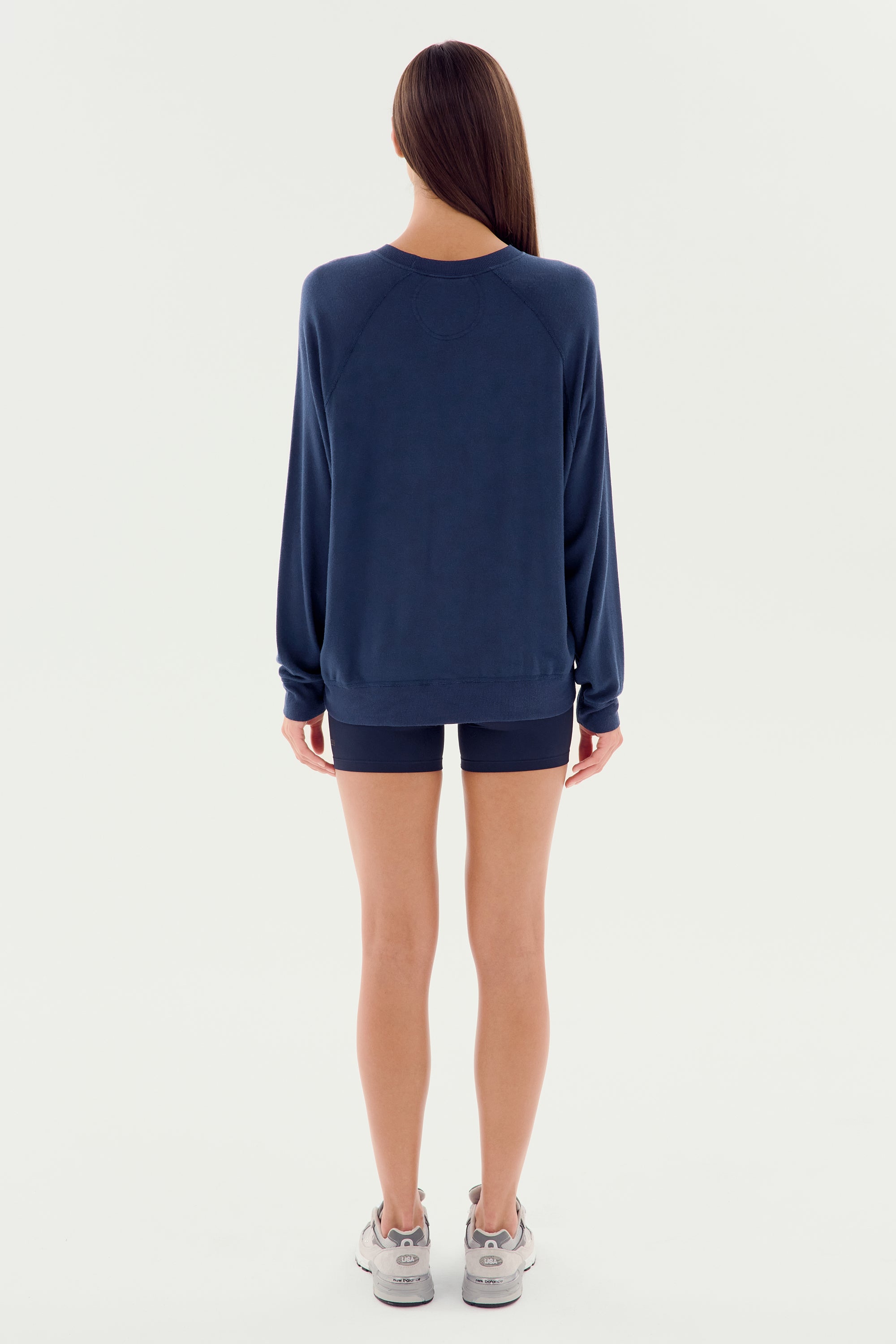 A person with long hair stands facing away, wearing the Andie Fleece Sweatshirt in Indigo from SPLITS59, matching navy blue shorts, and gray sneakers. The backdrop is a plain white. Garment proudly MADE IN LOS ANGELES.