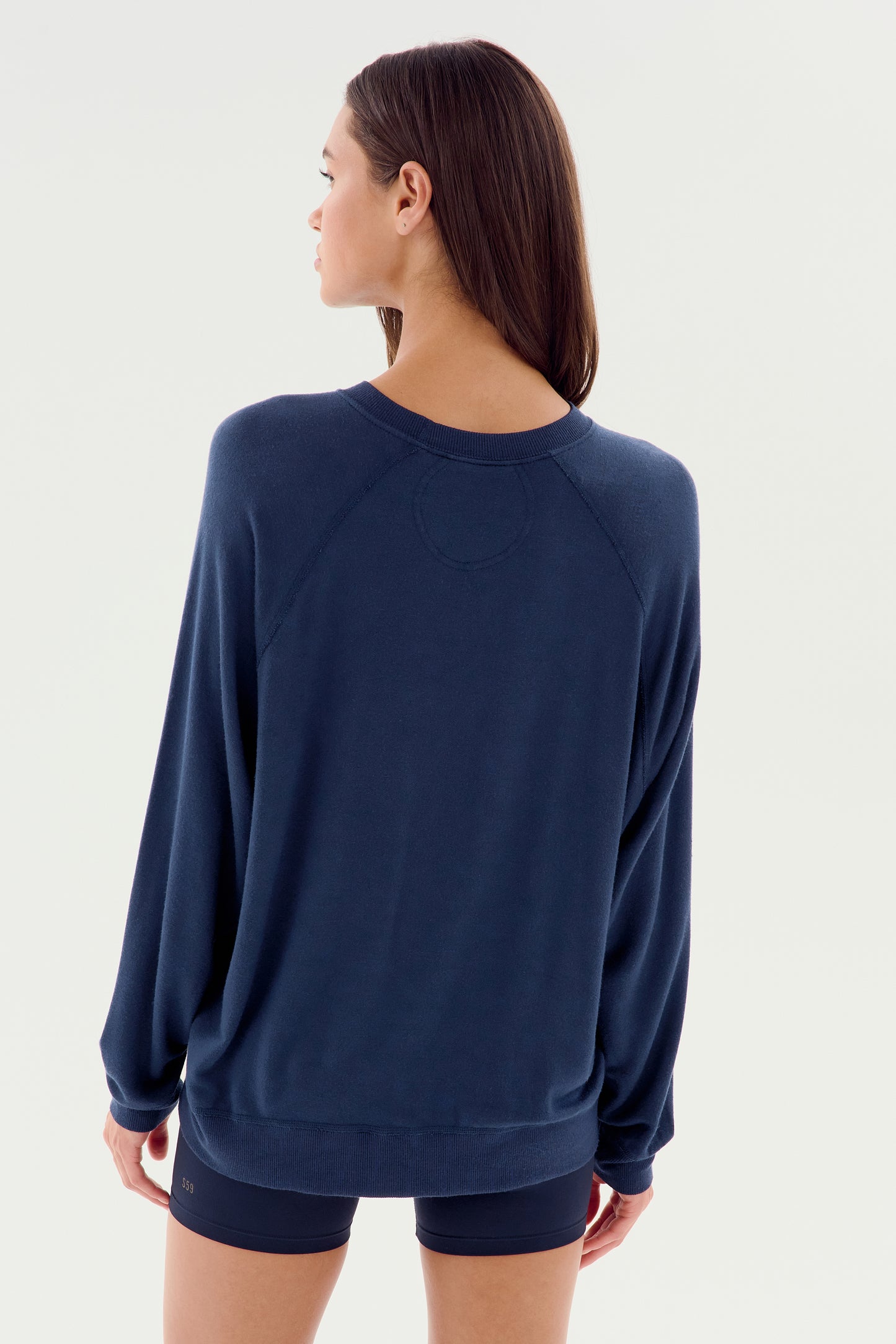 A person with long hair is seen from the back, wearing an SPLITS59 Andie Fleece Sweatshirt in Indigo and dark shorts, standing against a plain white background.