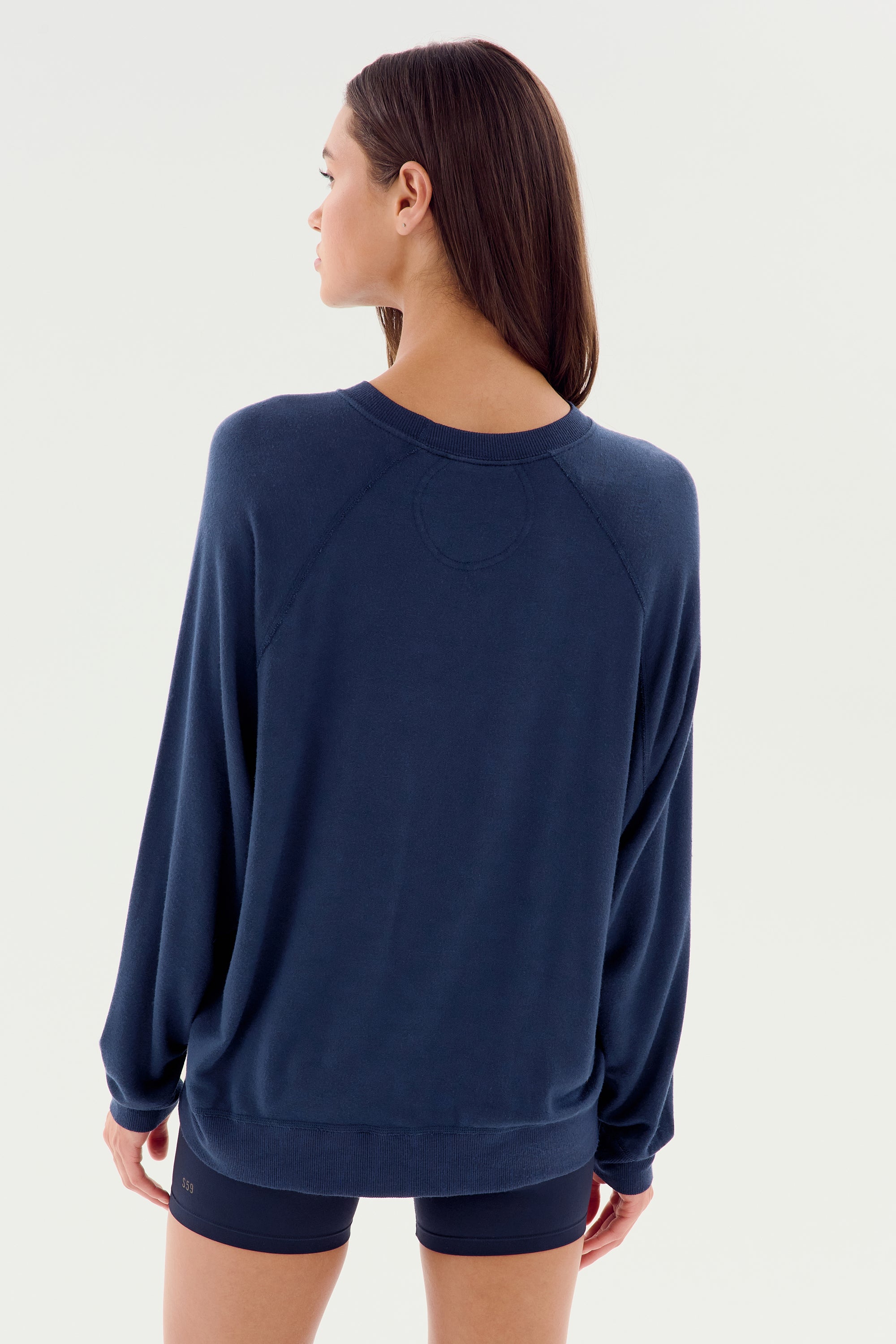 A person with long hair is seen from the back, wearing an SPLITS59 Andie Fleece Sweatshirt in Indigo and dark shorts, standing against a plain white background.