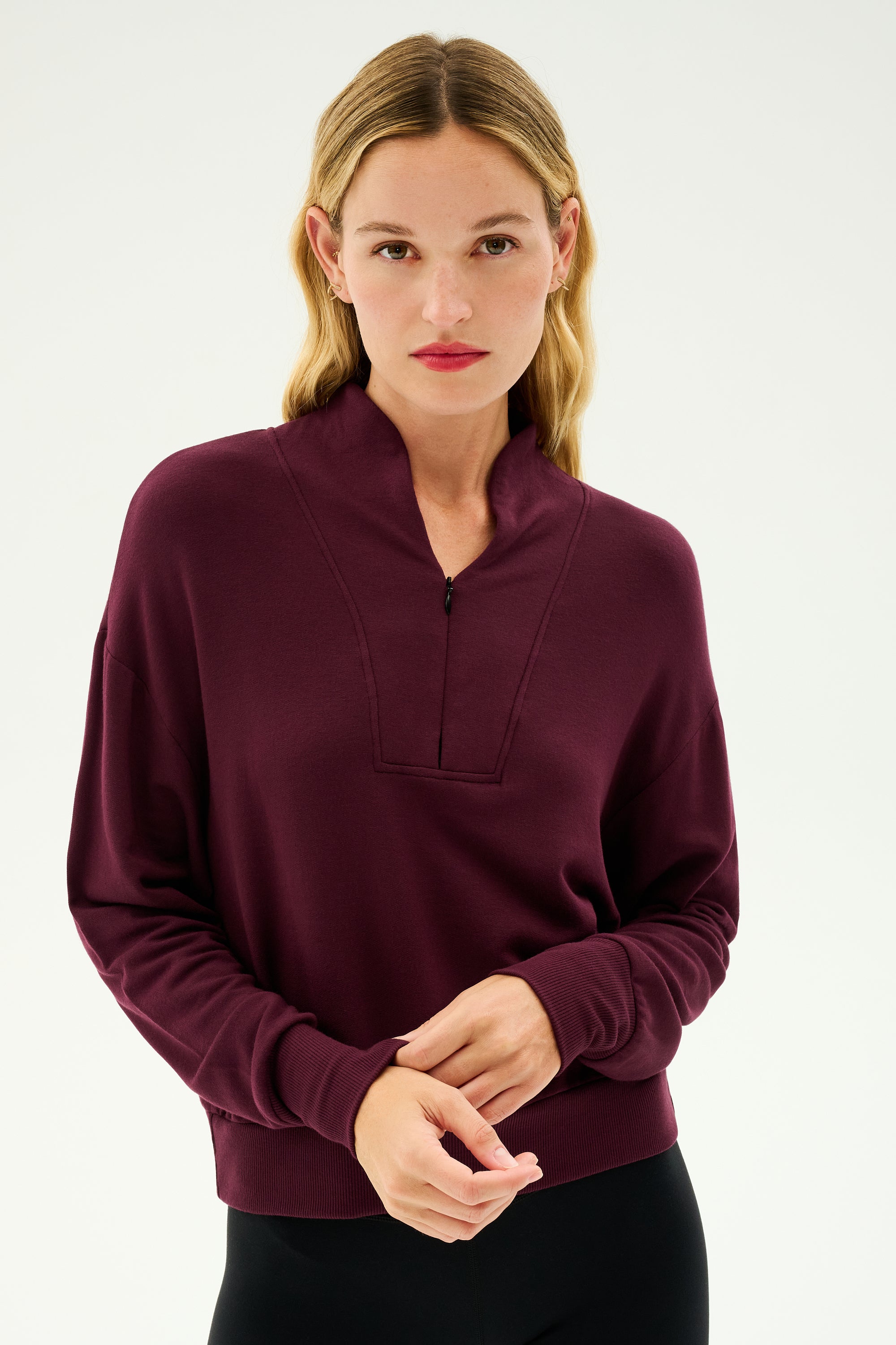 A person with long hair dressed in the cozy SPLITS59 Birdie Fleece Half Zip in Dark Cherry and black pants stands against a plain backdrop.