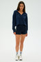 A woman in a navy Joan Fleece Polo Sweatshirt - Indigo by SPLITS59 and black shorts stands against a plain background, wearing white sneakers and socks.