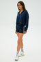 A person in an indigo Joan Fleece Polo Sweatshirt from SPLITS59 and athletic shorts with spandex stands against a plain background, wearing white socks and sneakers.