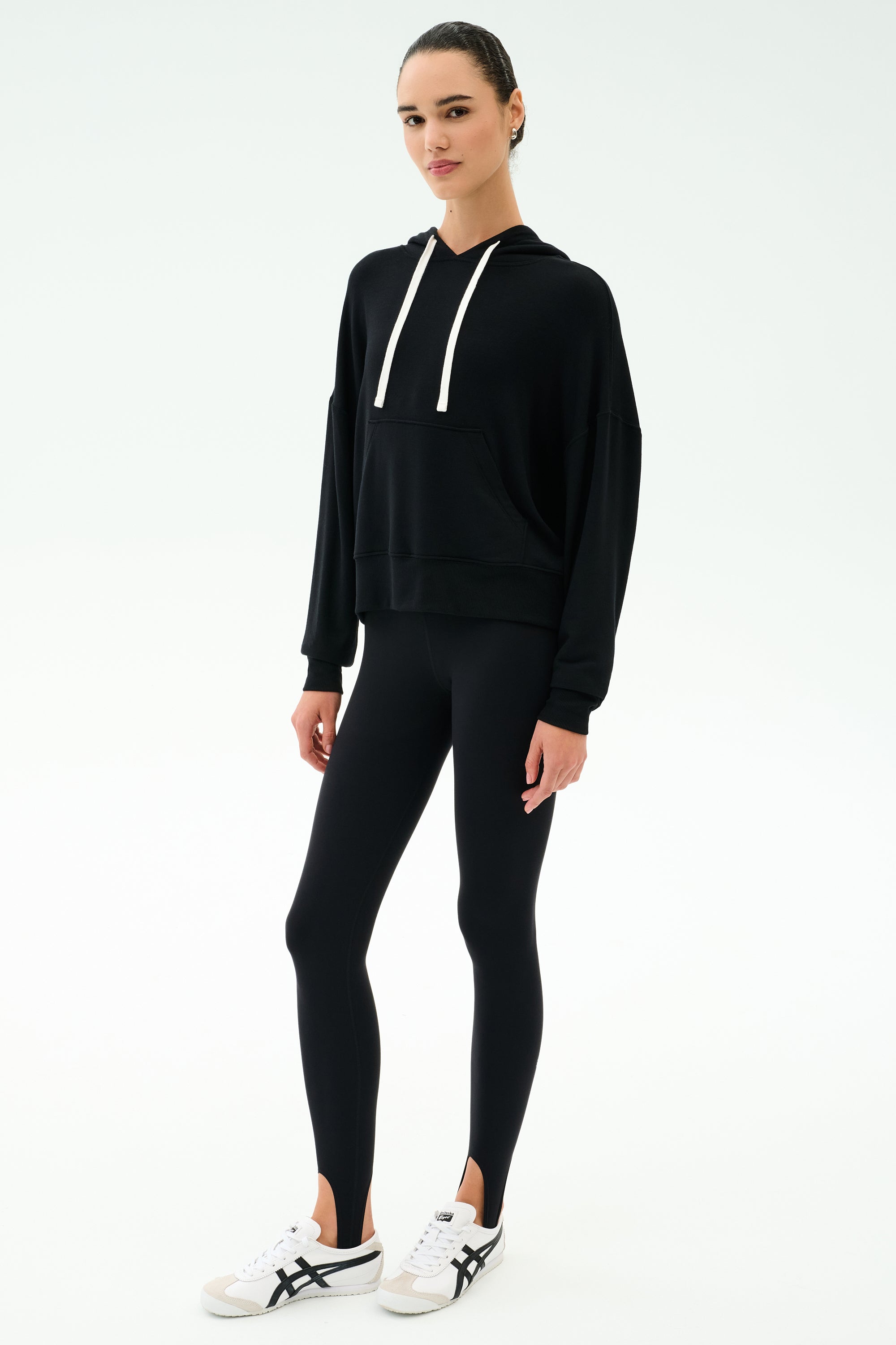 A woman faces forward against a plain white background, wearing the SPLITS59 Joey Long Sleeve Hoodie in black, paired with black leggings and white sneakers featuring black accents.