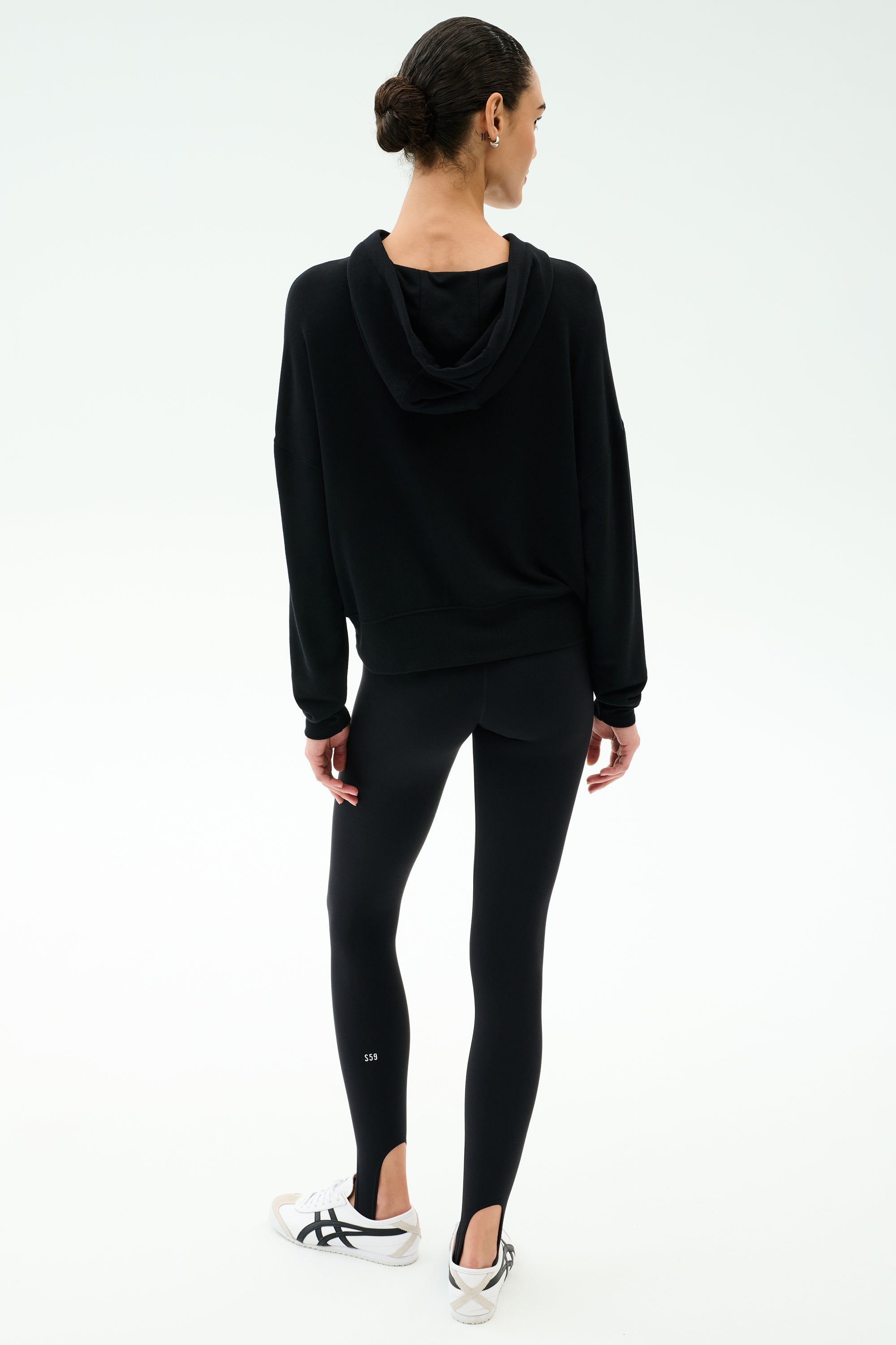 A person stands facing away, wearing the SPLITS59 Joey Long Sleeve Hoodie in black, paired with black leggings and white sneakers against a plain white background.