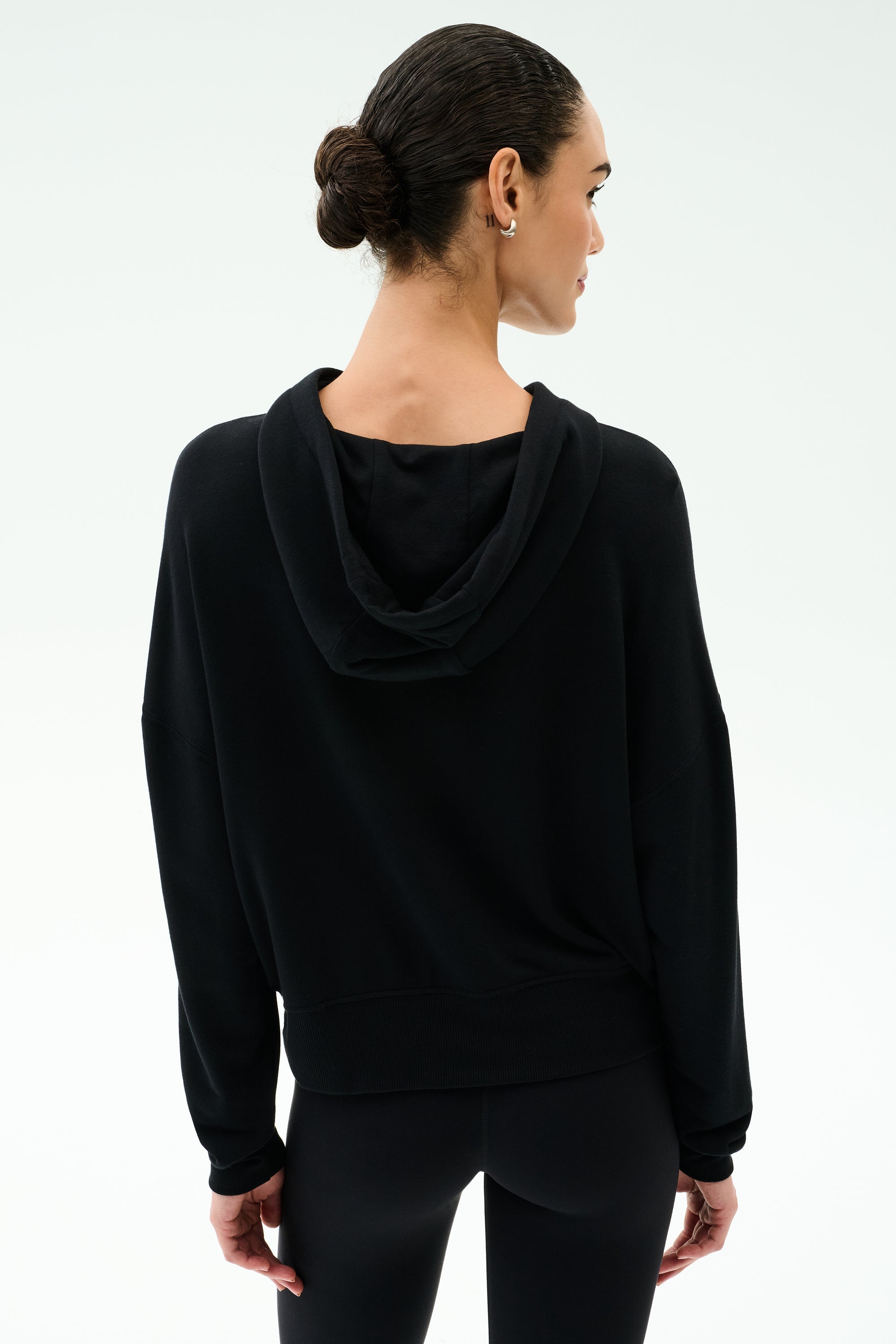 A person with dark hair in a bun wears the SPLITS59 Joey Long Sleeve Hoodie in black and black spandex-blend leggings, facing away from the camera against a plain light background.