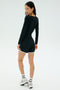 A woman stands looking over her shoulder, wearing SPLITS59's Megan Woven Nylon Run Short in black, a black long-sleeve spandex top, white socks, and silver sneakers against a plain background.
