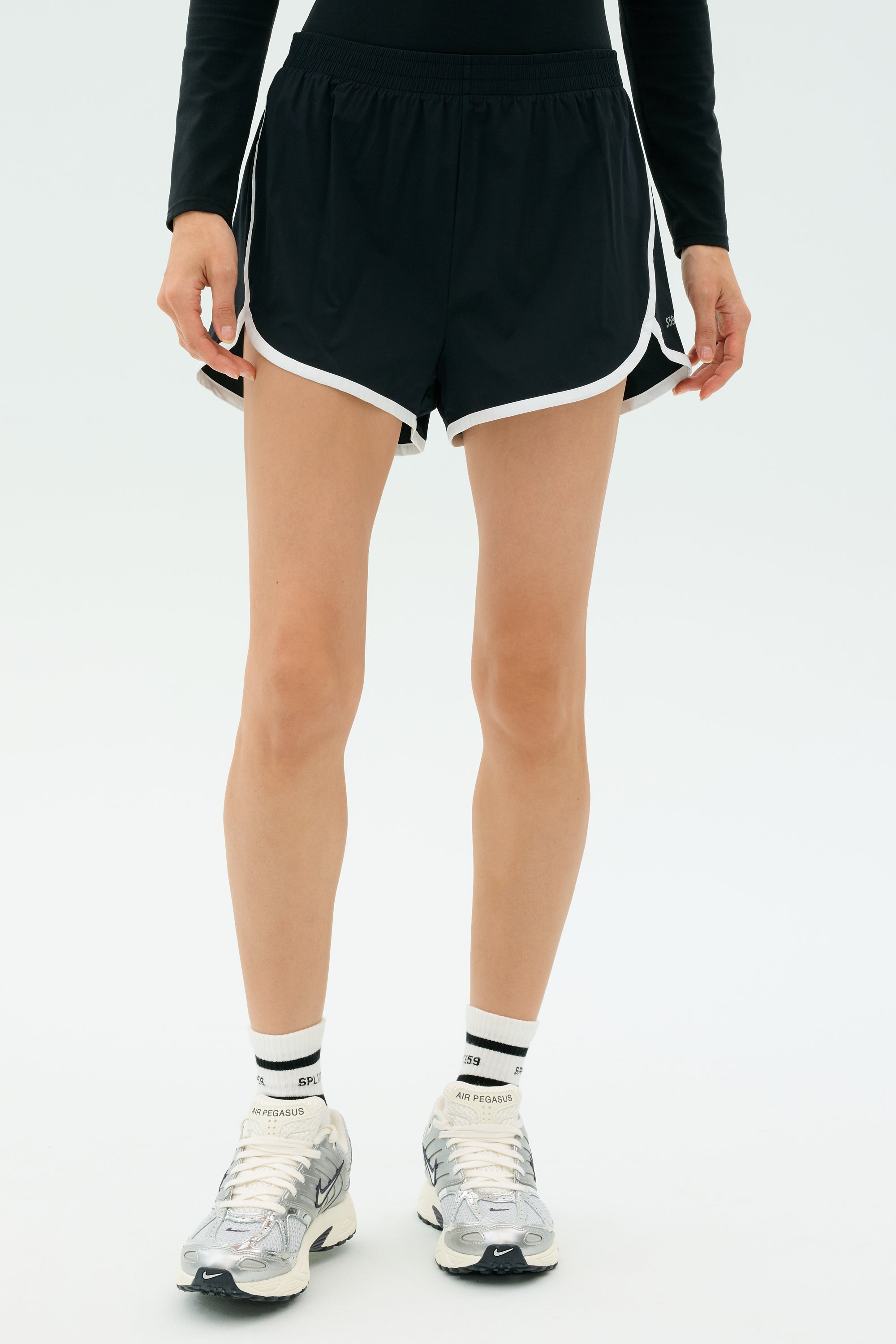 A person wears SPLITS59 Megan Woven Nylon Run Shorts in black with white trim, paired with white socks and silver running shoes, standing against a light background.