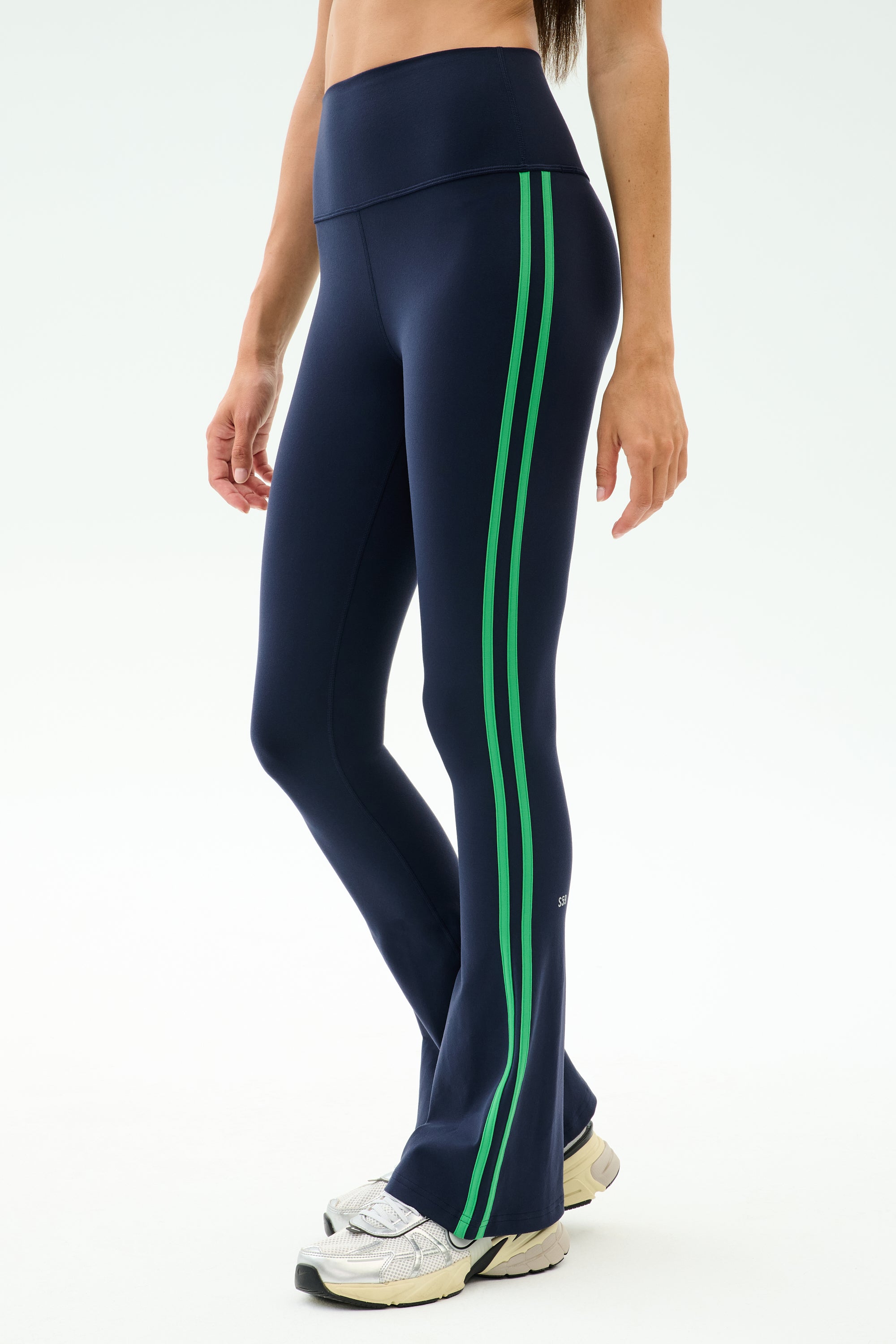 A person wears SPLITS59 Raquel High Waist Supplex Flare leggings in Indigo/Grass—navy with two green side stripes—paired with white sneakers, shown waist to feet. These 4-way stretch leggings are ideal for workouts or casual wear.