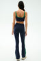 A woman faces away, wearing SPLITS59’s Raquel High Waist Supplex Flare in Indigo/Grass with a matching sports bra and white sneakers, set against a plain white background.