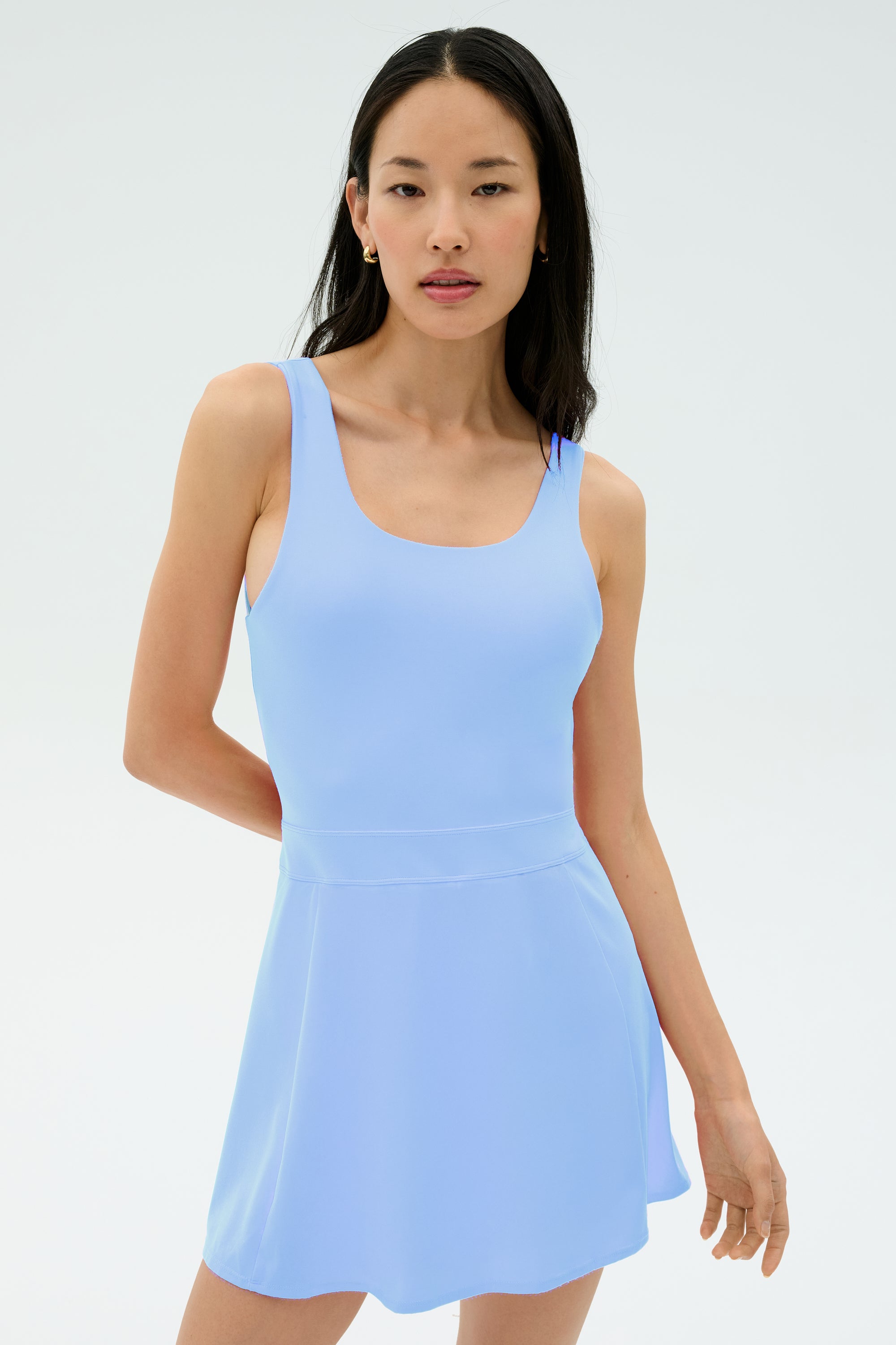 A woman wears the SPLITS59 Martina Rigor Dress - Skylight, a light blue sleeveless style, standing against a white background and looking at the camera—a versatile pick for court sports or casual outings.