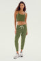 A woman stands wearing a green crop top made from feminine drape stretch modal fabric, complemented by the Olive Reena Fleece 7/8 Sweatpants from SPLITS59 and white sneakers, all set against a neutral background.