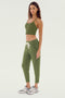 A woman stands against a plain white background dressed in an olive cropped tank top paired with the SPLITS59 Reena Fleece 7/8 Sweatpant - Olive, and grey sneakers. The ensemble's feminine drape stretch modal fabric ensures both comfort and style.