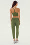 A woman is standing with her back to the camera, wearing a green sports bra, SPLITS59 Reena Fleece 7/8 Sweatpant in Olive made of feminine drape stretch modal fabric, and grey sneakers against a plain background.