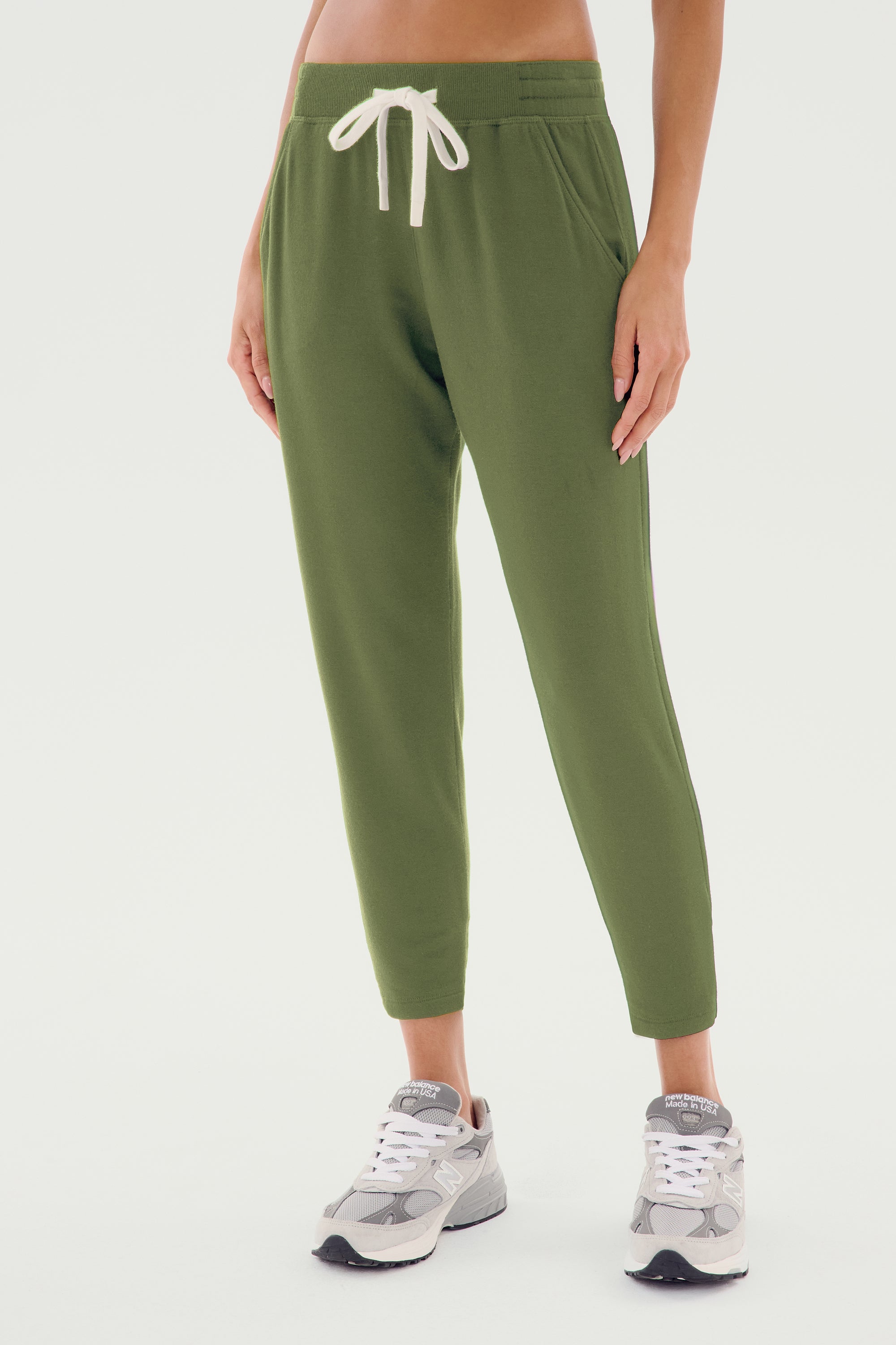 A person is wearing the SPLITS59 Reena Fleece 7/8 Sweatpant in Olive with an elastic waistband and white drawstring, paired with gray sneakers and a classic rugby polo.