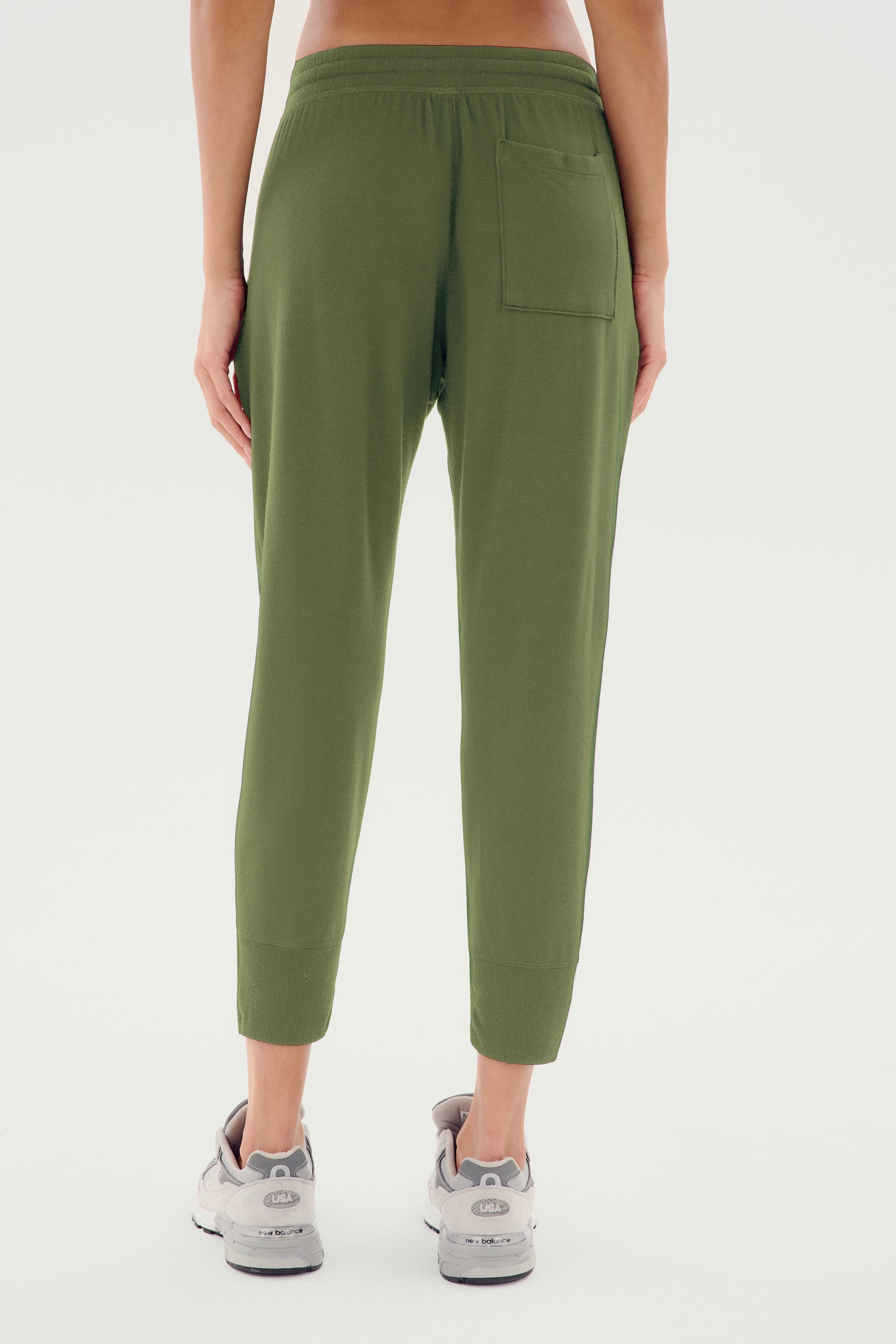 A person is standing facing away, wearing the SPLITS59 Reena Fleece 7/8 Sweatpant in olive and gray sneakers. Made from a feminine drape stretch modal fabric, these sweatpants feature a back pocket on the right side.
