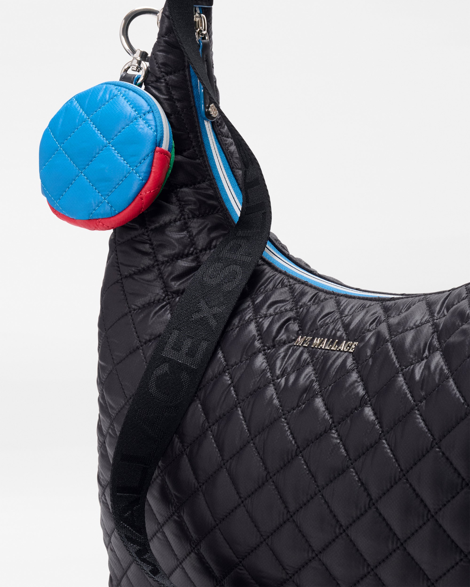 Close-up of the MZW x Splits59 Carry All by SPLITS59—a black quilted bag with logo, thick black strap, blue zipper, and a small round blue and red quilted pouch attached.