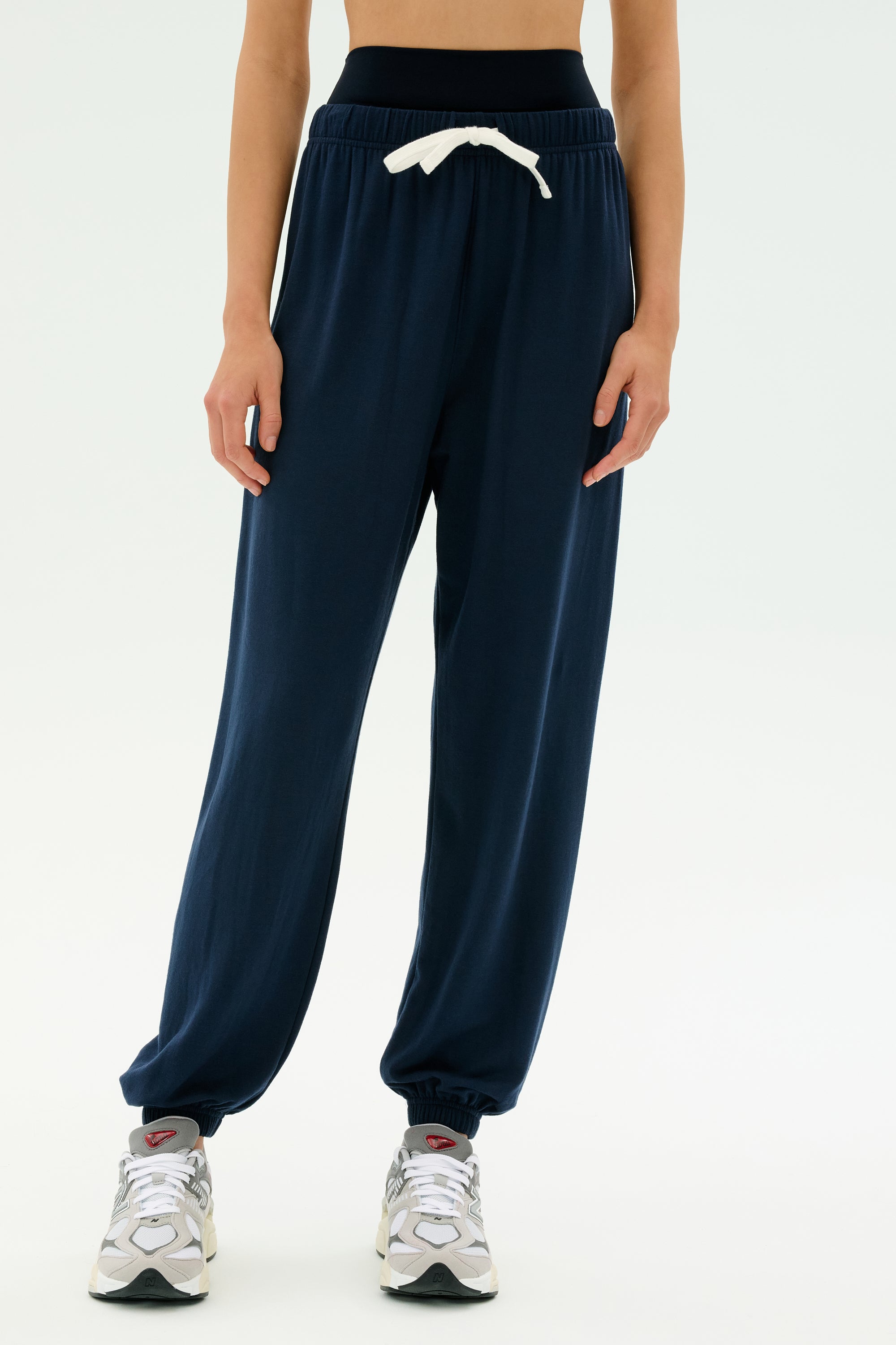 A person wears SPLITS59’s Andie Oversized Fleece Sweatpant in Indigo with a white drawstring, gray sneakers, and a black crop top against a white background—ideal for cool weather workouts. MADE IN LOS ANGELES.