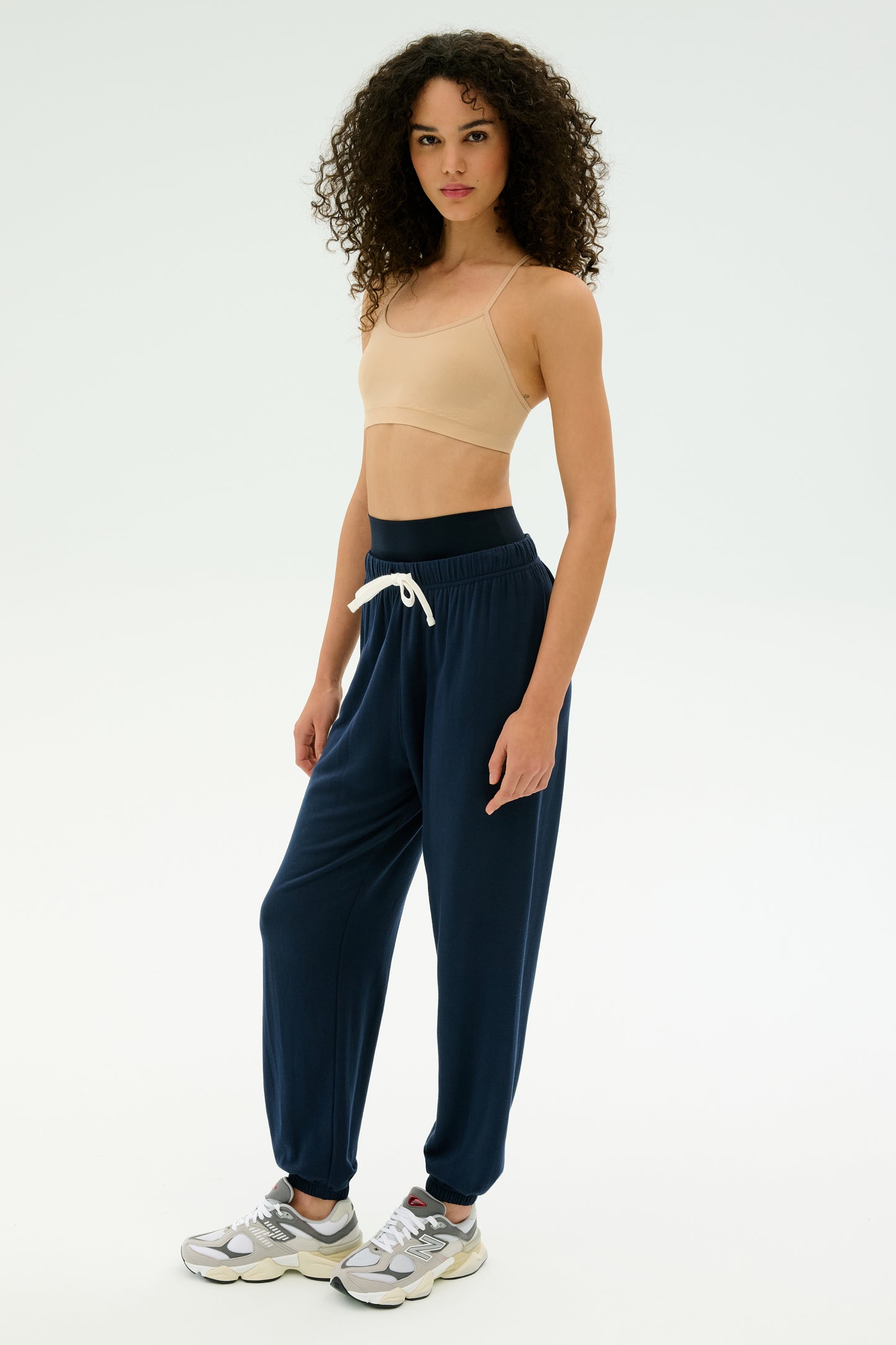 A person with curly hair wears a beige crop top, SPLITS59 Andie Oversized Fleece Sweatpant in Indigo, and silver sneakers, standing against a plain light background—perfect for cool weather workouts.