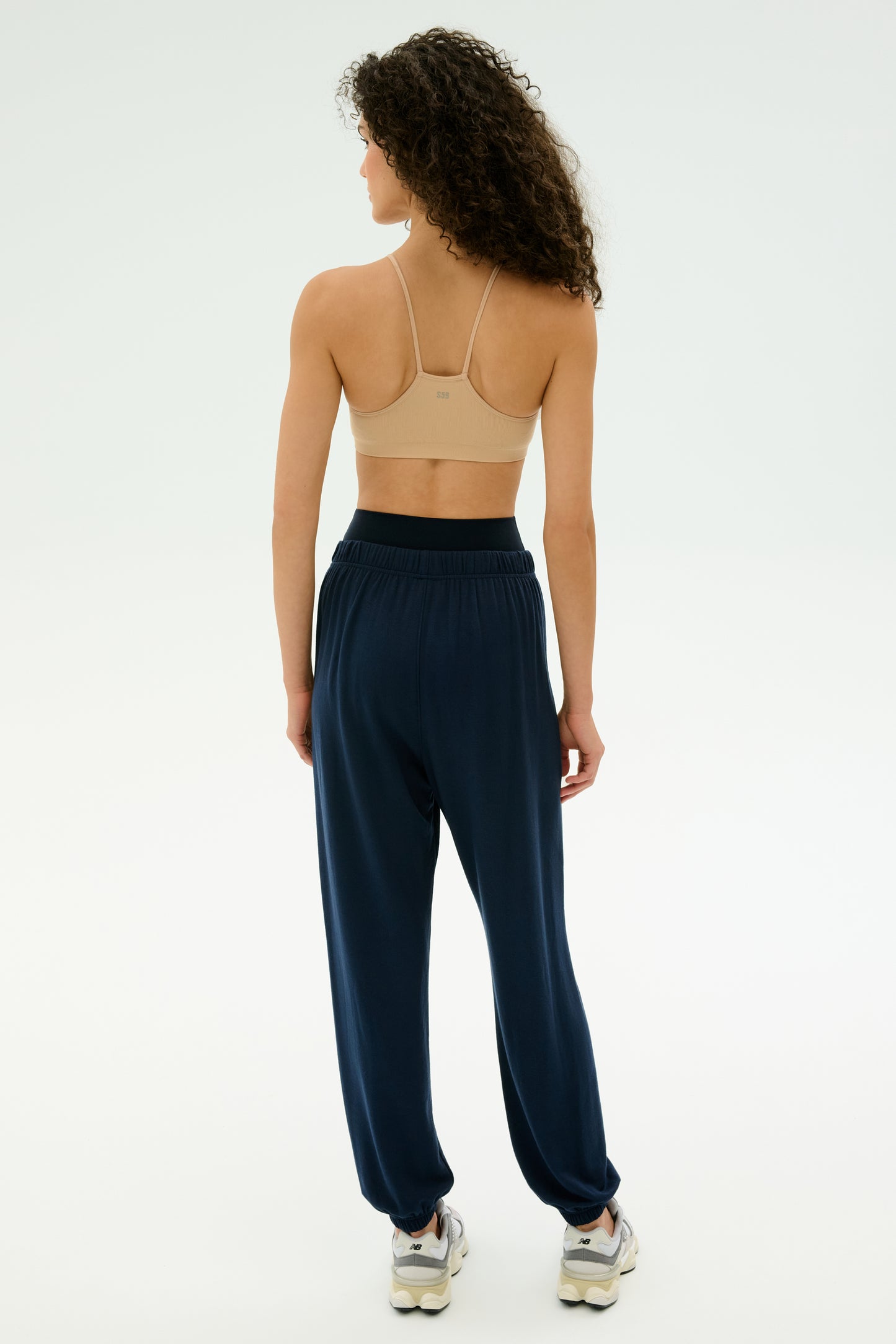 A woman with curly hair faces away, wearing a beige sports bra, SPLITS59 Andie Oversized Fleece Sweatpant in Indigo, and white sneakers against a plain light background—ideal for cool weather workouts.