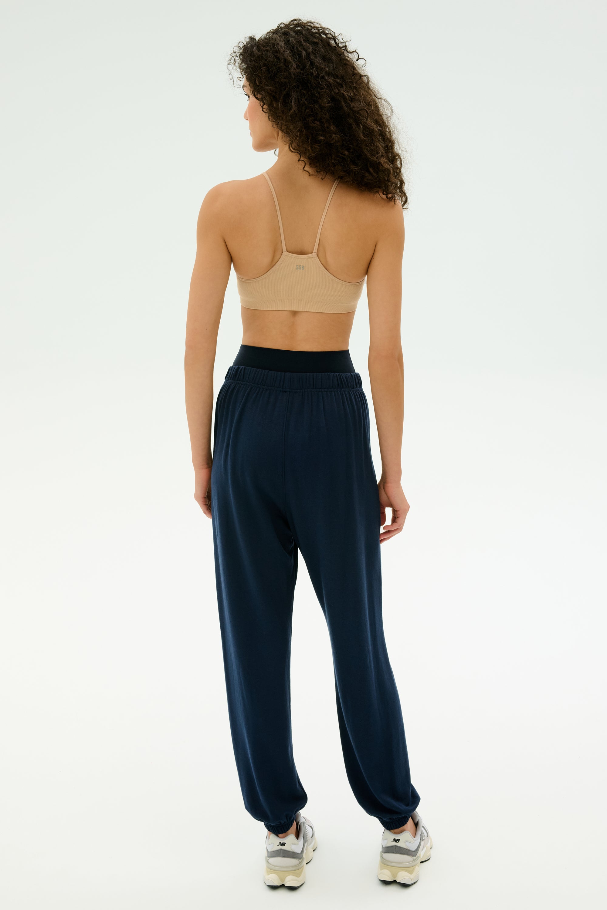 A woman with curly hair faces away, wearing a beige sports bra, SPLITS59 Andie Oversized Fleece Sweatpant in Indigo, and white sneakers against a plain light background—ideal for cool weather workouts.