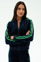 A woman with long dark hair poses in SPLITS59's Heritage Velour Bomber in Indigo/Grass, featuring green stripes—perfect for yoga warmups—against a plain white background.