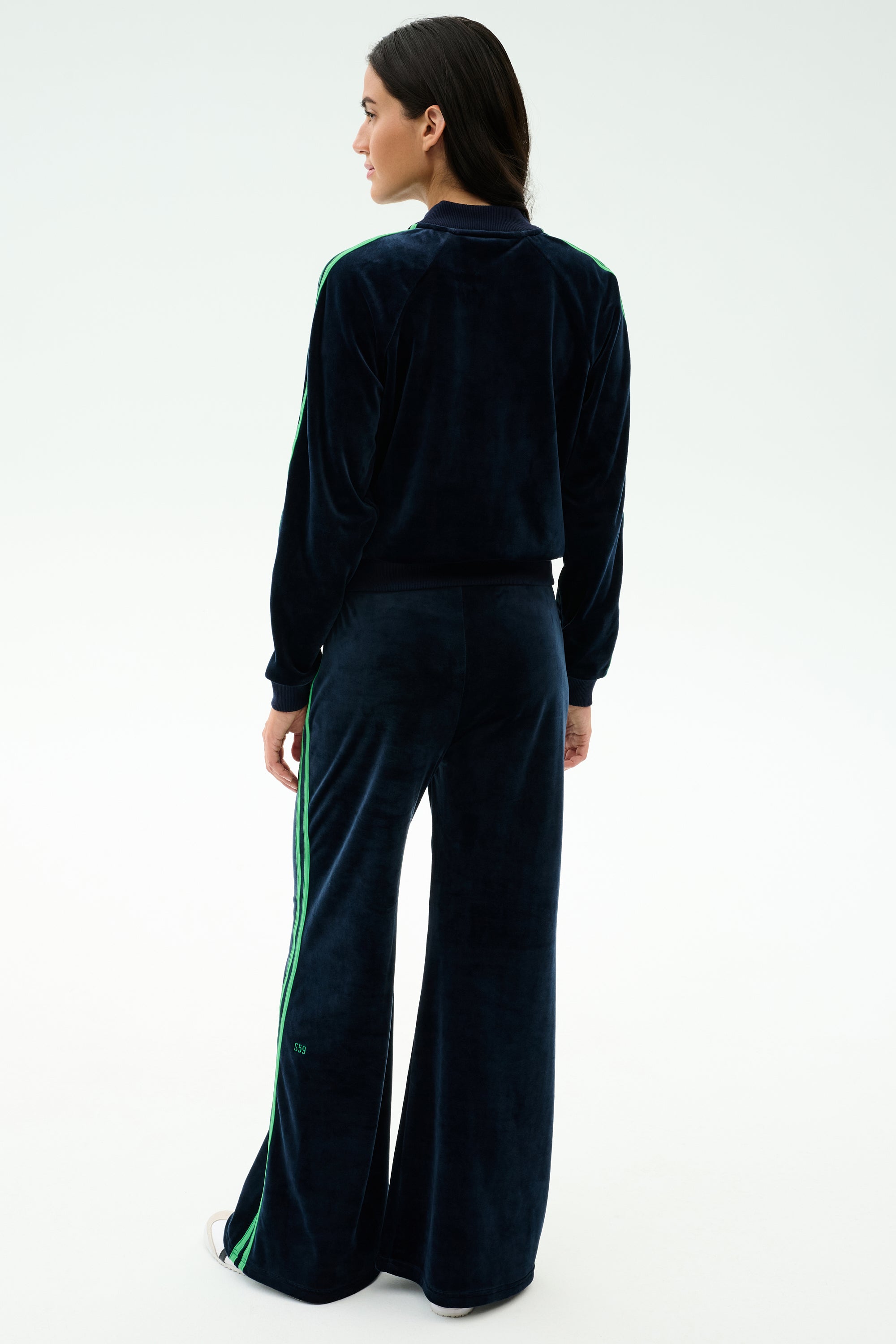 A person stands with their back to the camera wearing the SPLITS59 Heritage Velour Bomber in Indigo/Grass, featuring green stripes along the arms and legs, paired with white shoes against a plain light background.