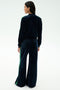 A person stands with their back to the camera wearing the SPLITS59 Heritage Velour Bomber in Indigo/Grass, featuring green stripes along the arms and legs, paired with white shoes against a plain light background.