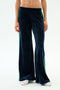 A person models the SPLITS59 Heritage Velour Pant in Indigo/Grass, featuring wide-leg navy blue velour fabric with green side stripes, ankles visible above white sneakers against a plain white background.