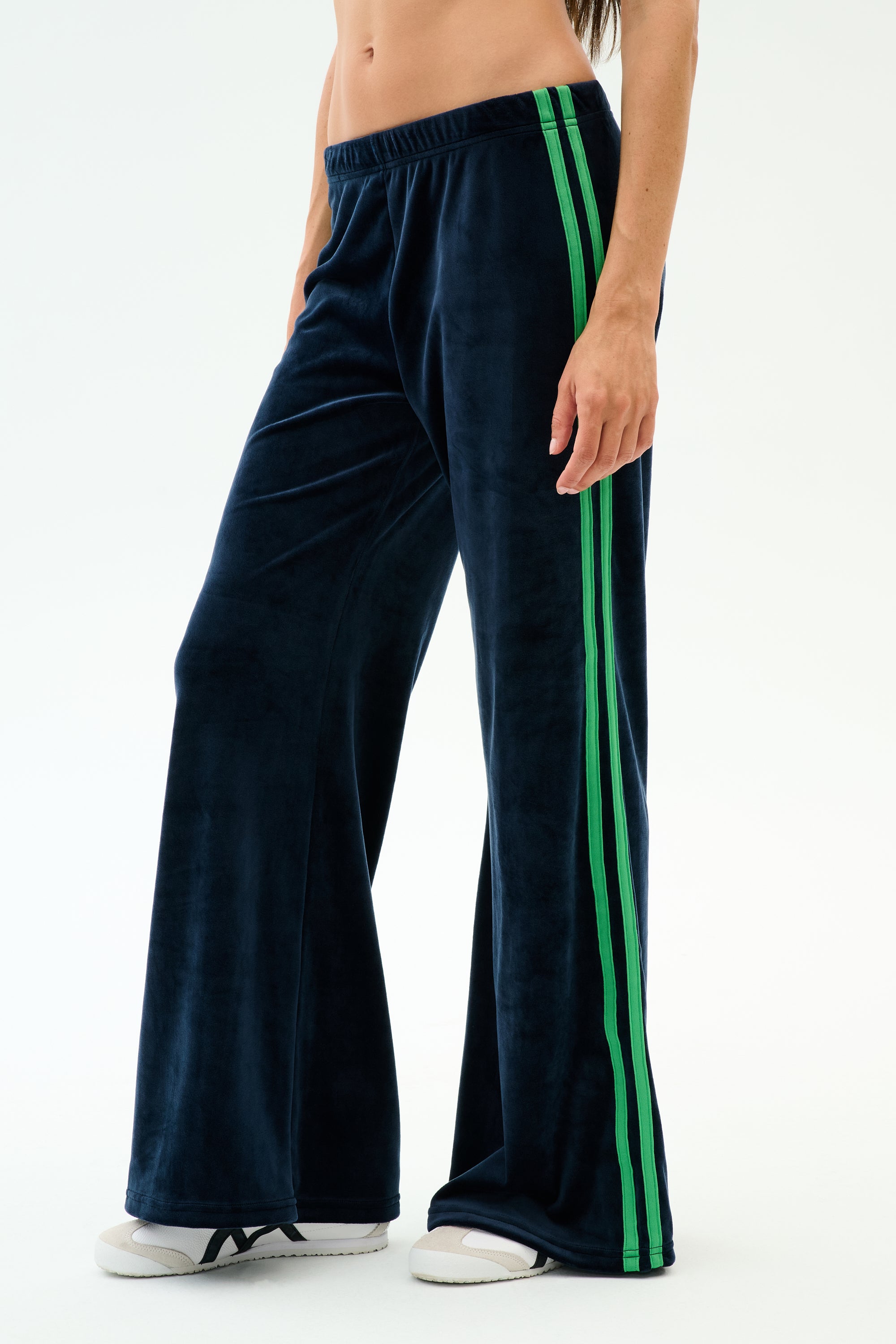 A person models the SPLITS59 Heritage Velour Pant in Indigo/Grass, featuring green side stripes, paired with white sneakers against a plain white background.
