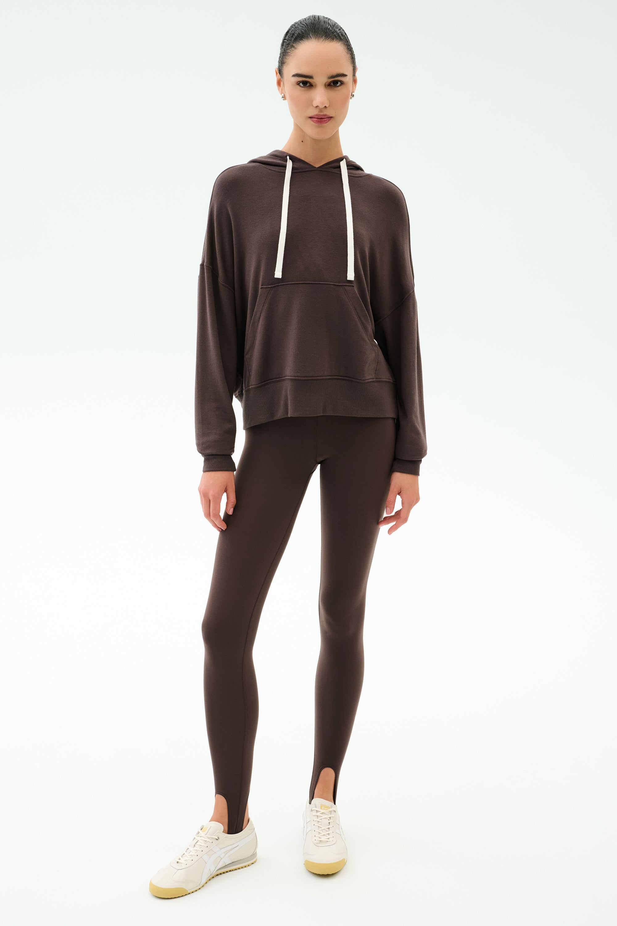 A person stands facing forward in the SPLITS59 Joey Long Sleeve Hoodie - Dark Chocolate and matching brown leggings, both in a soft modal spandex blend, paired with white sneakers against a plain white background.