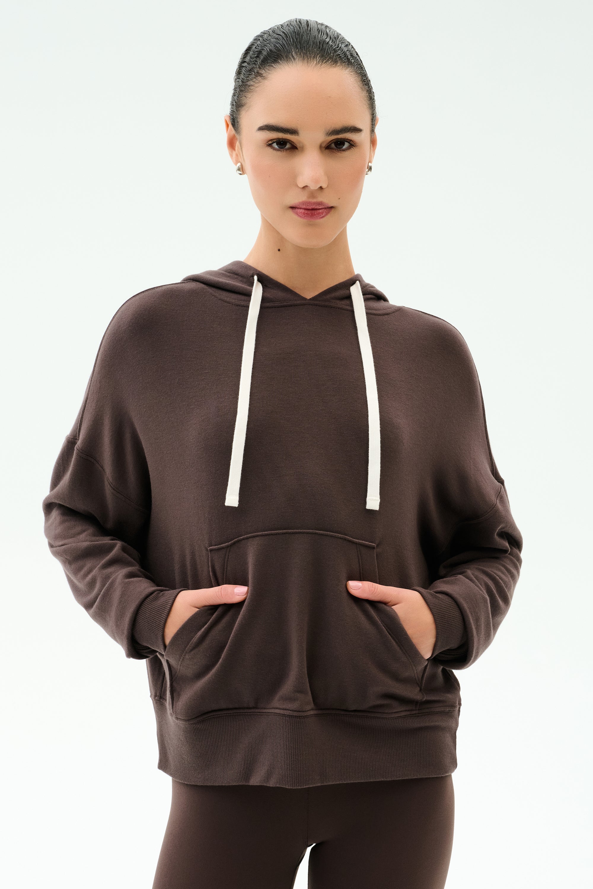 A person wears the SPLITS59 Joey Long Sleeve Hoodie in Dark Chocolate with matching pants, hands in the hoodie’s front pocket, facing the camera against a plain light background.