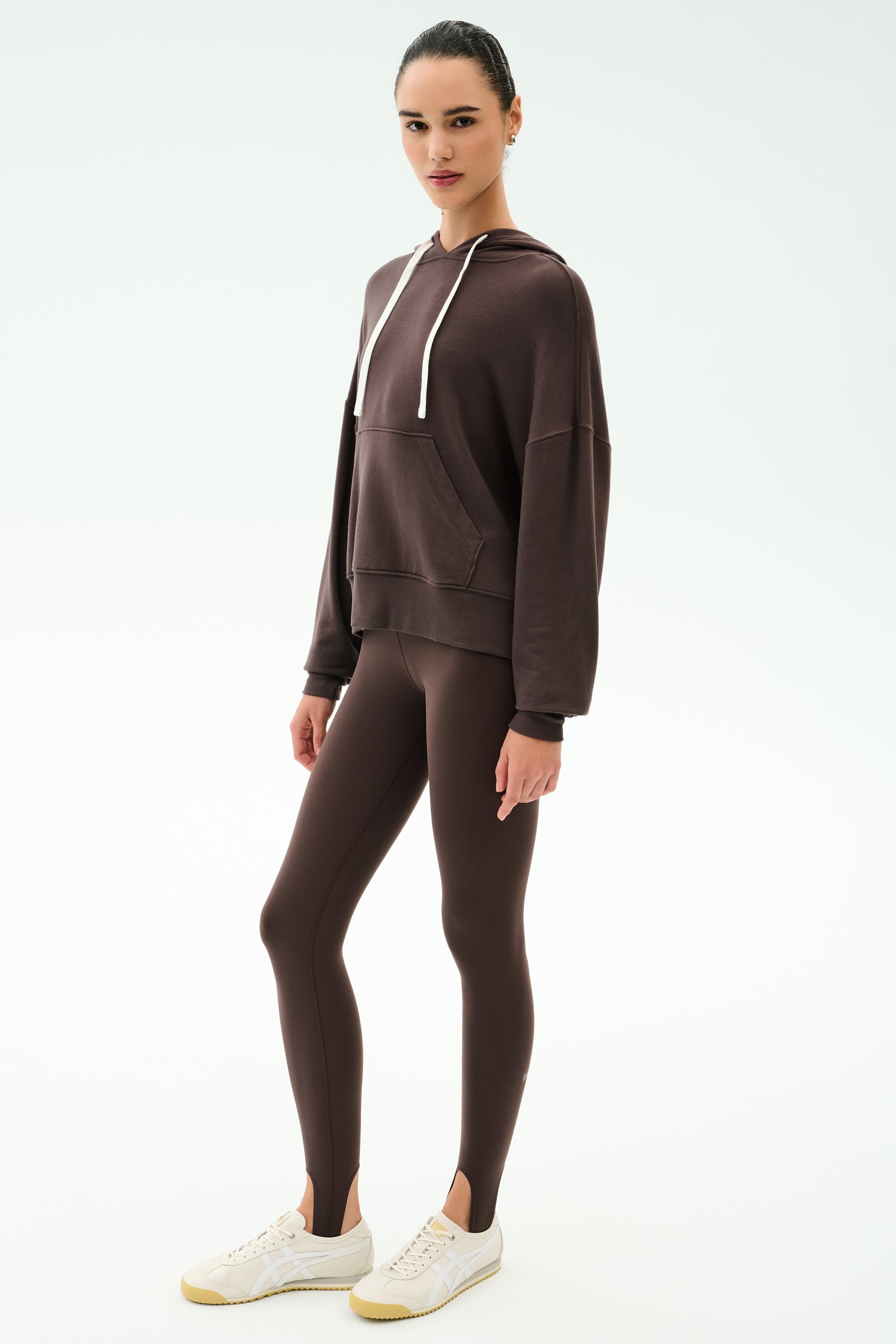 A woman stands on a white background wearing the SPLITS59 Joey Long Sleeve Hoodie in Dark Chocolate, matching brown leggings with stirrups, and white sneakers.