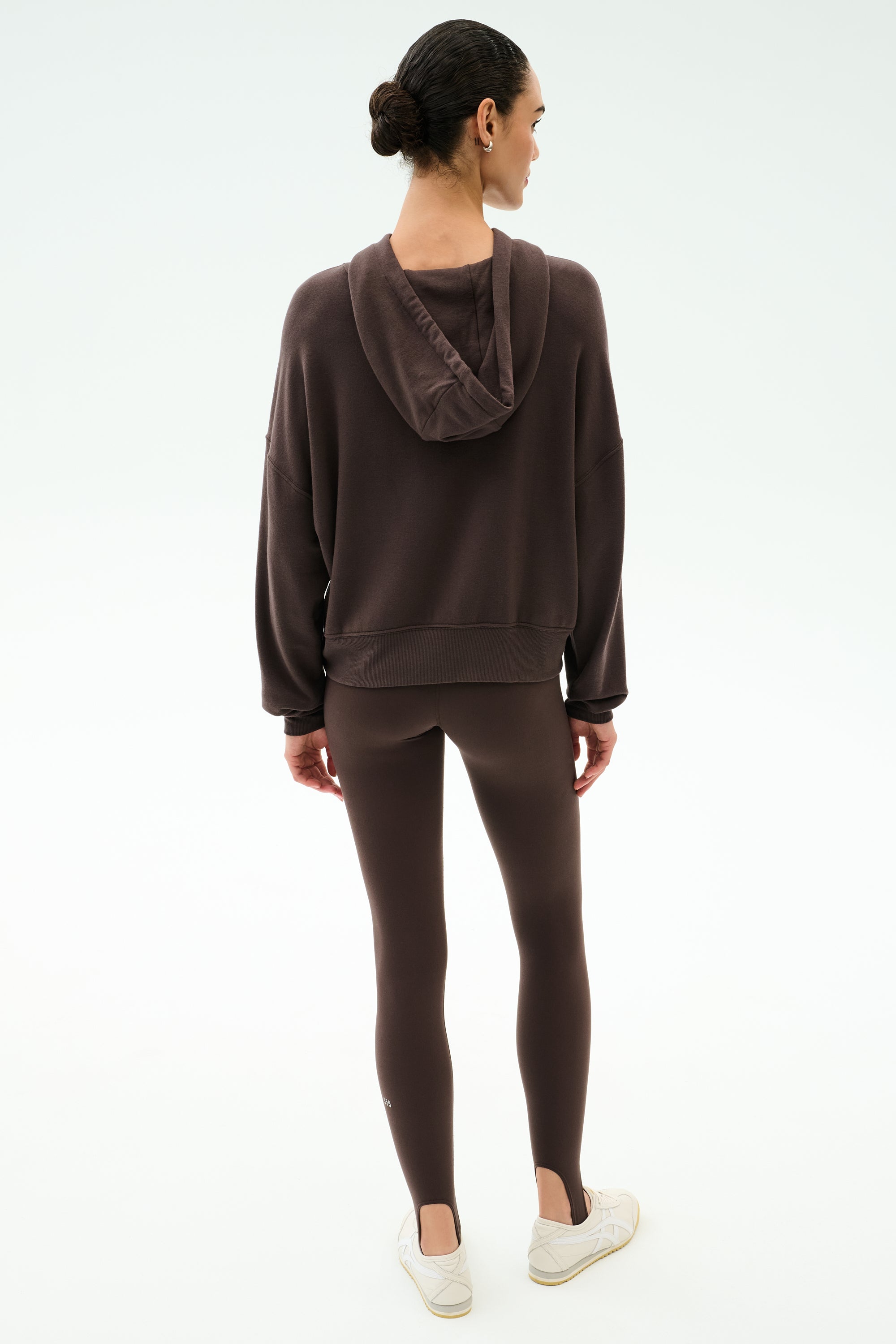 A person stands facing away, wearing the SPLITS59 Joey Long Sleeve Hoodie in Dark Chocolate, brown leggings, and white sneakers against a plain light background.