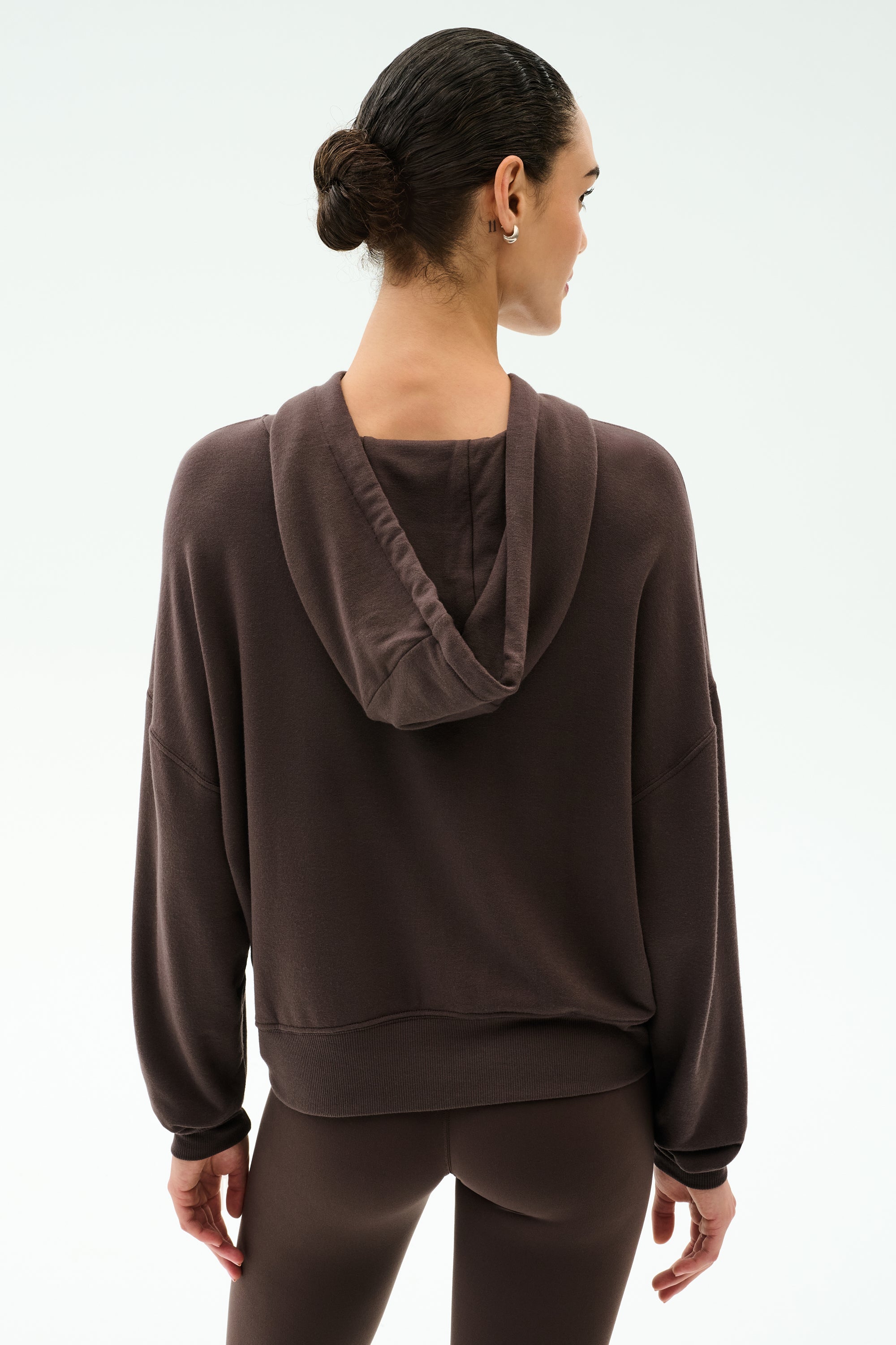 A person with dark hair in a low bun is seen from the back wearing the SPLITS59 Joey Long Sleeve Hoodie in Dark Chocolate with matching spandex blend leggings, set against a plain light background.