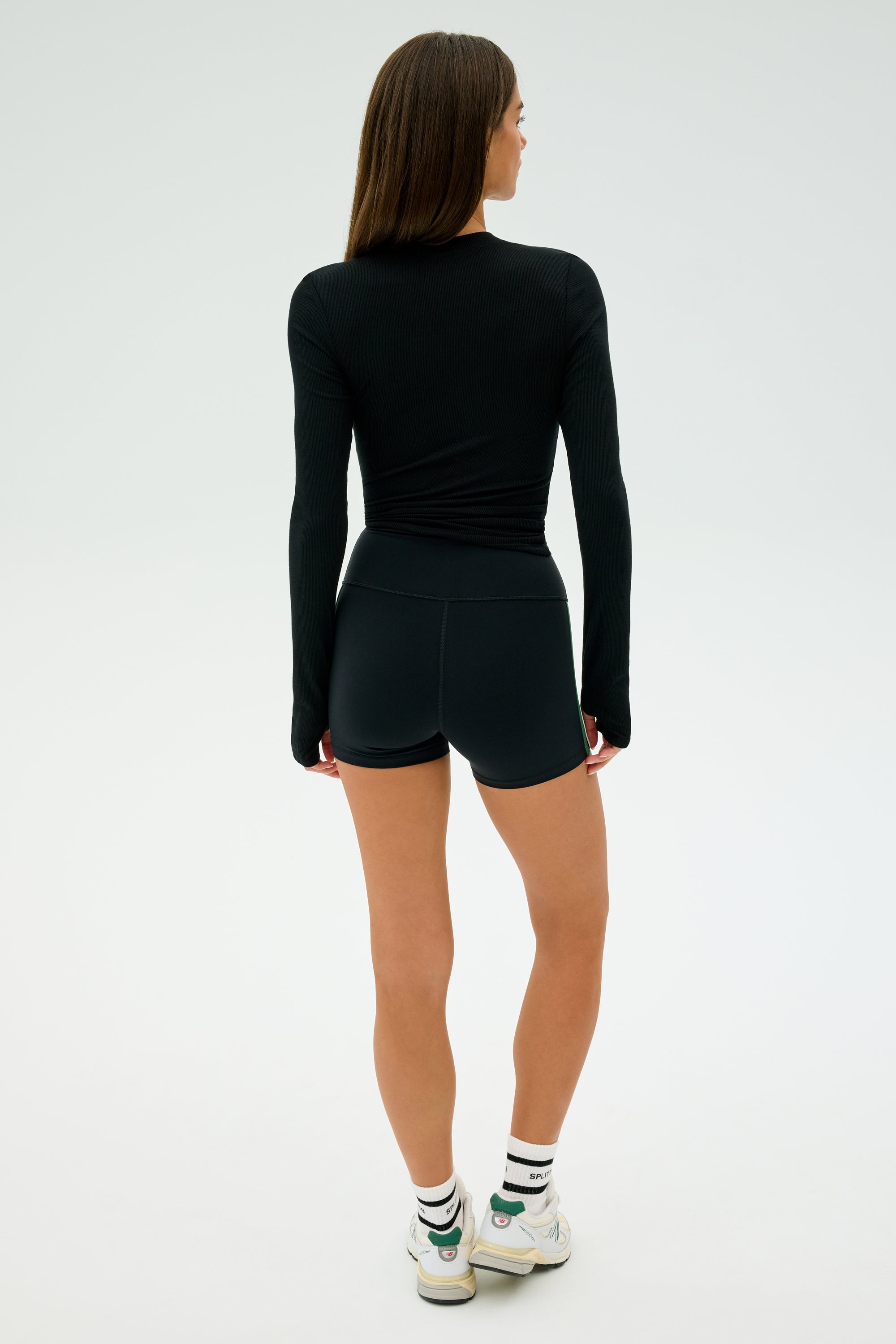 A woman stands facing away in SPLITS59's Ella Airweight High Waist Short 3.5" in Black/Arugula, styled with a black long-sleeve top, white sneakers, and black-striped socks against a plain white background.